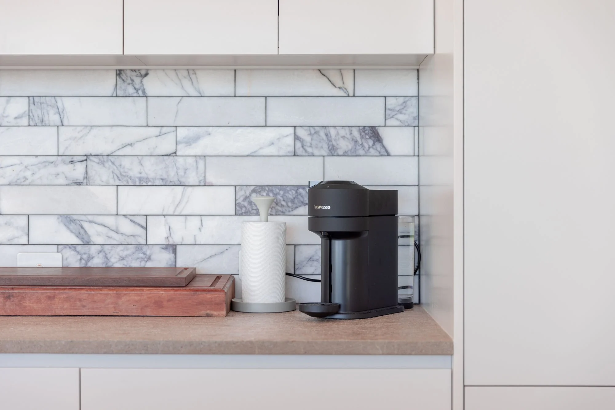 A kitchen countertop with a black espresso machine, a white roll of paper towels, and a small white bowl on a paper towel holder against a white marble tile backsplash.