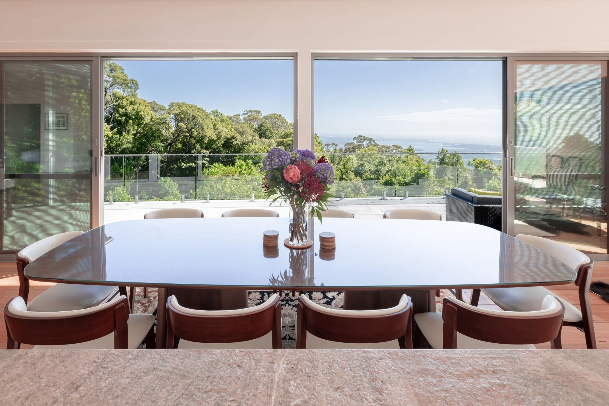 A dining area with a glass-top table surrounded by white and wooden chairs. On the table, there is a glass vase with a bouquet of purple, red, and pink flowers. The room has large sliding glass doors that open to a balcony with a glass railing, overl