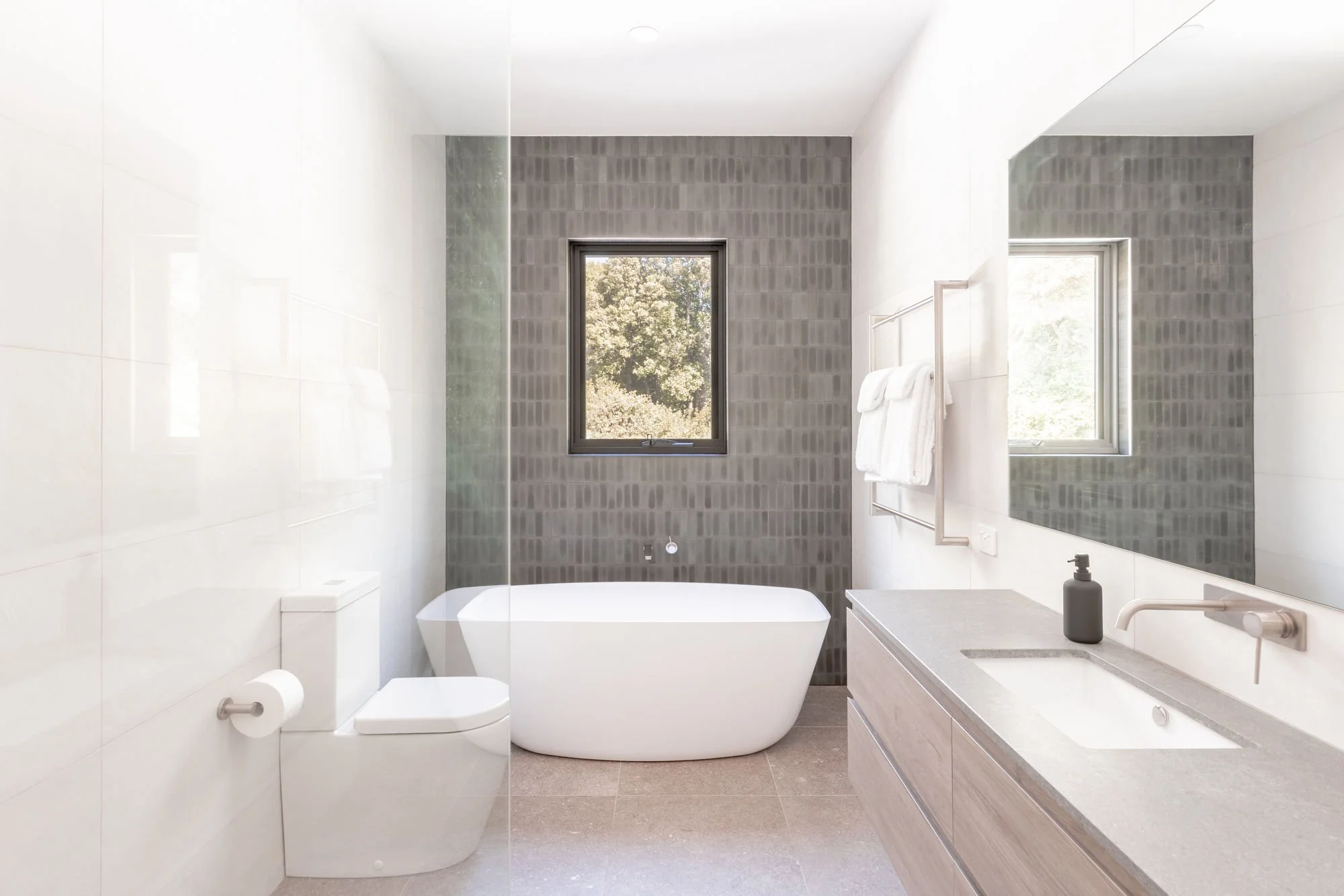 Modern bathroom with white freestanding bathtub, large windows, gray tiled accent wall, white vanity with sink, soap dispenser, and mirror, with beige tiled floor.