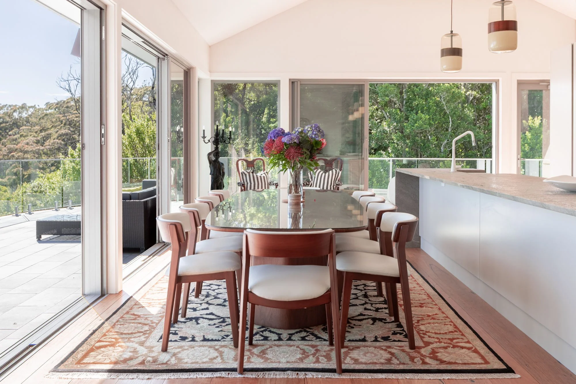 A bright, modern dining room with a large wooden dining table surrounded by white chairs, a vase of colorful flowers, a patterned rug, and large glass sliding doors that open to an outdoor patio with seating and lush green trees.