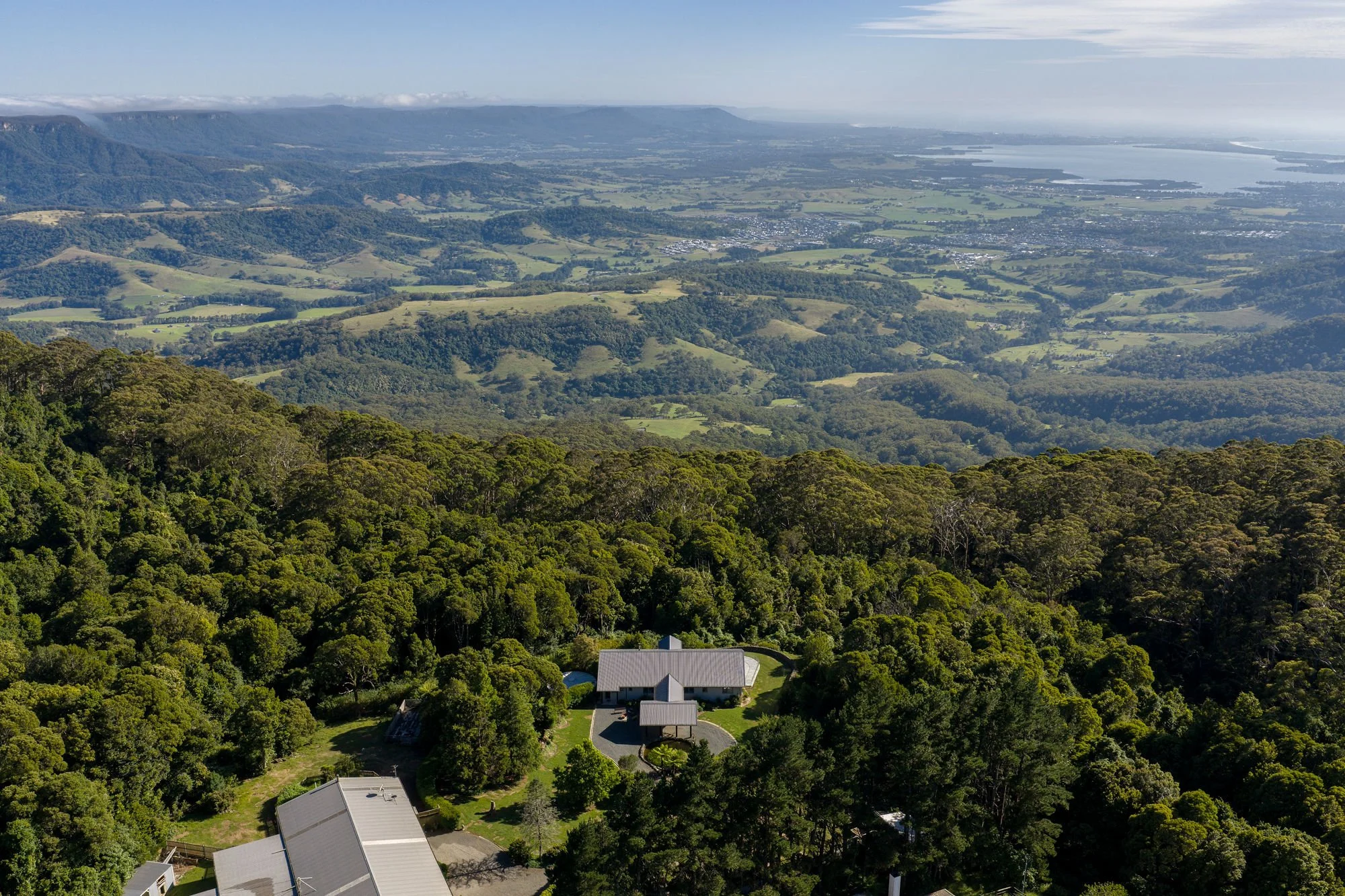 Aerial view of a lush green landscape with trees, houses, and rolling hills, with a lake and distant mountains under a partly cloudy sky.