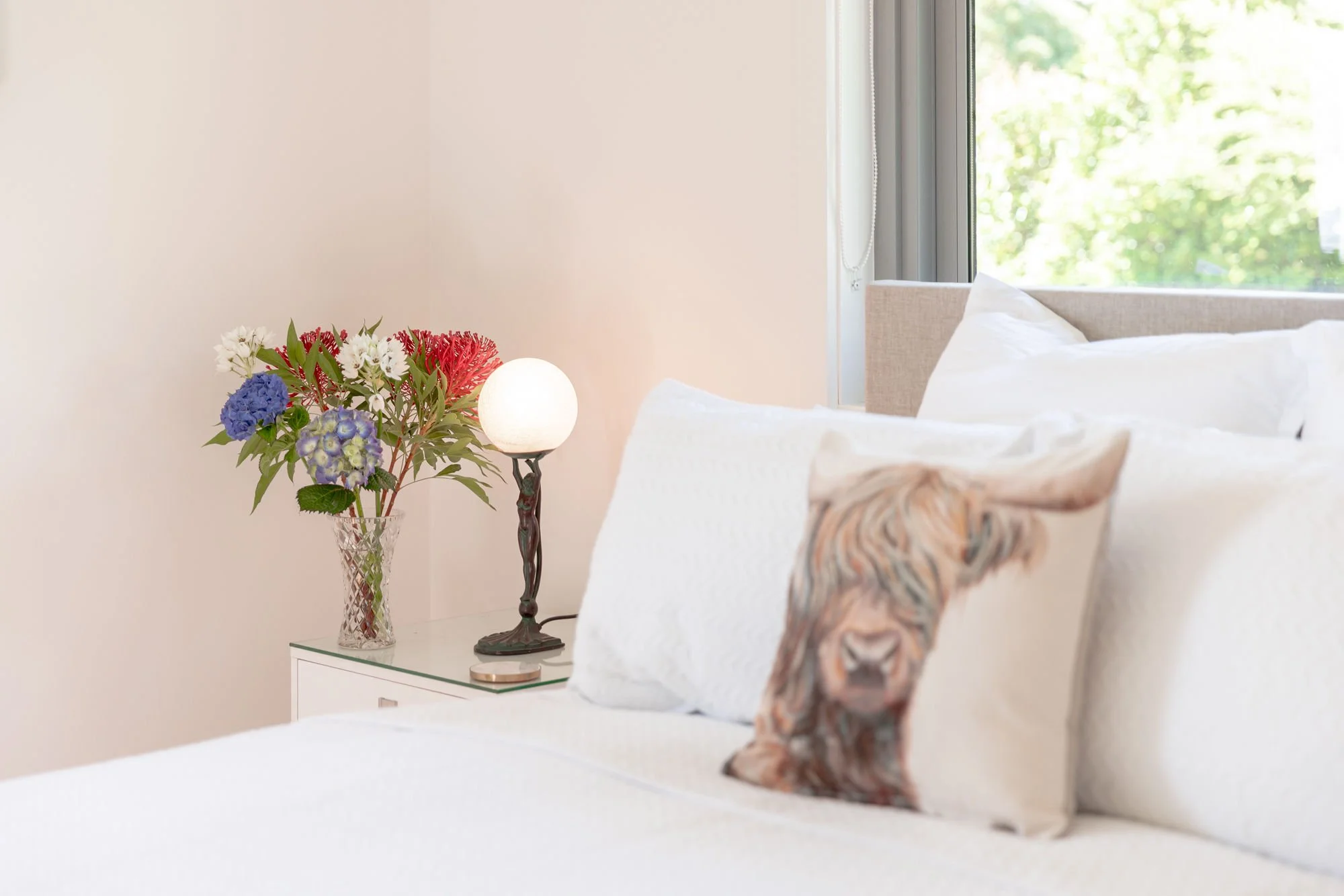 A bedroom with a bedside table, a vase of colorful flowers, a lamp, a bed with white bedding, and a window with green trees outside.