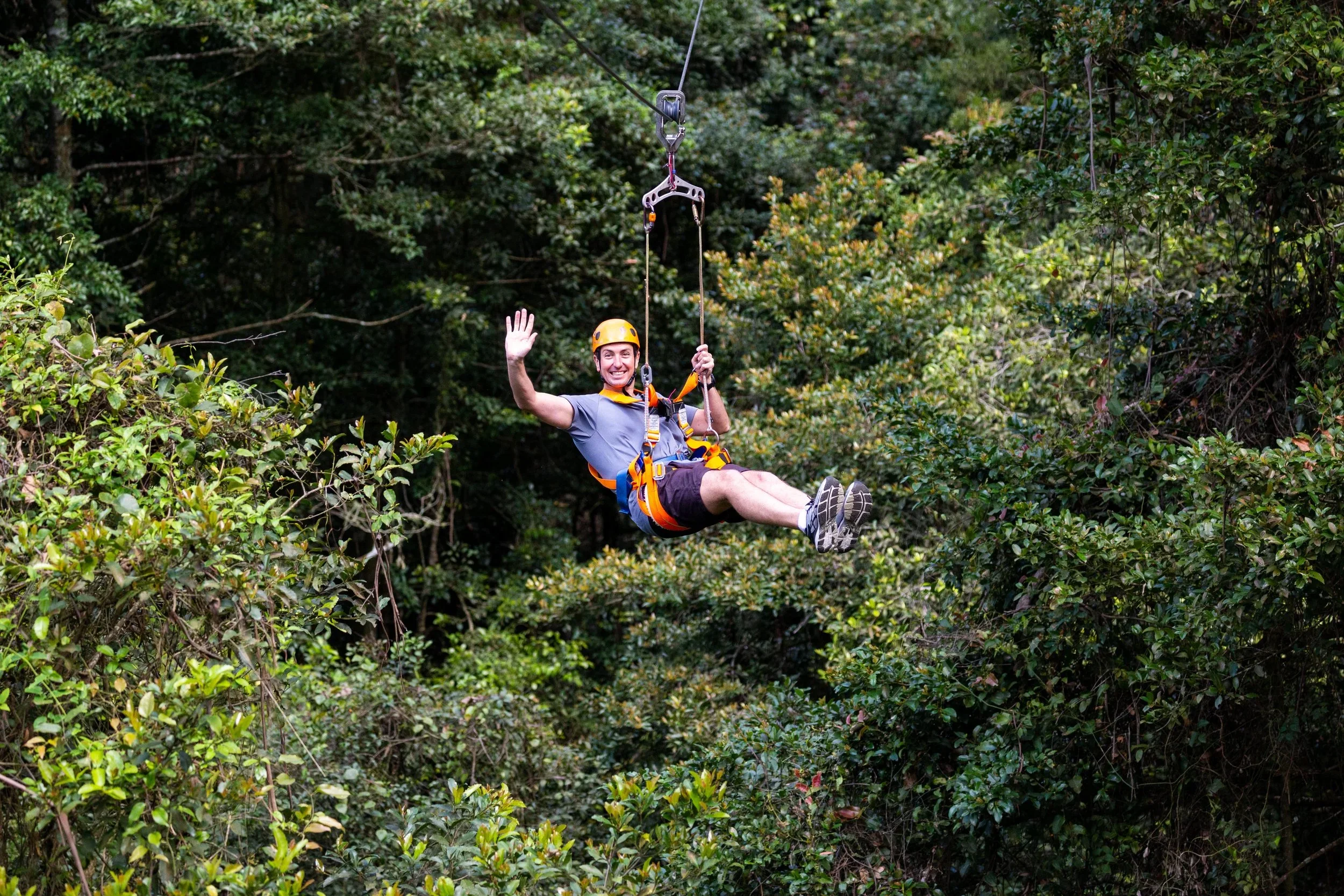 A man wearing a yellow helmet and gray t-shirt ziplining through a wooded area, smiling and waving at the camera.