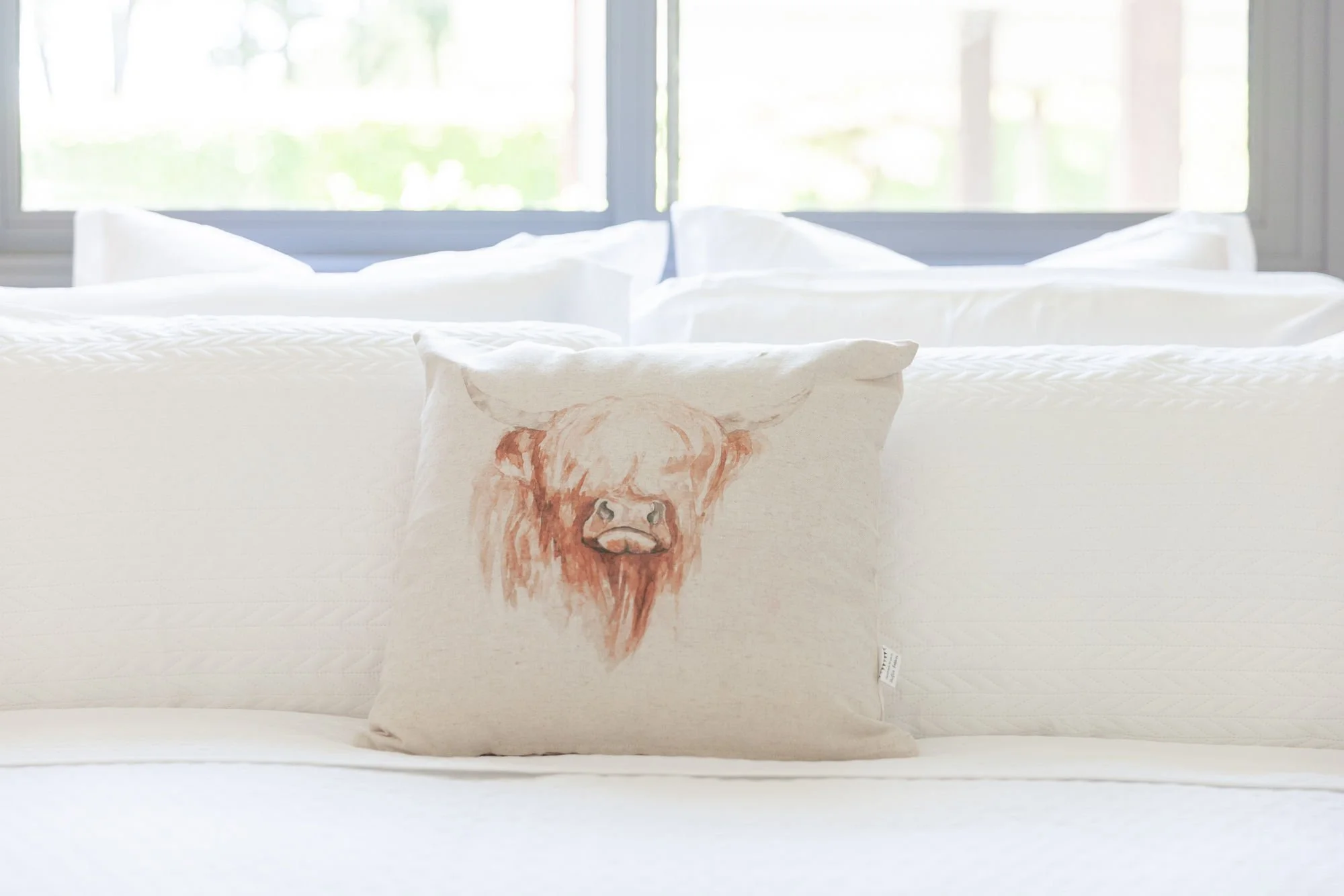 Cream-colored bed with a decorative pillow featuring a lion's face, situated in a brightly lit room with large windows and trees outside.