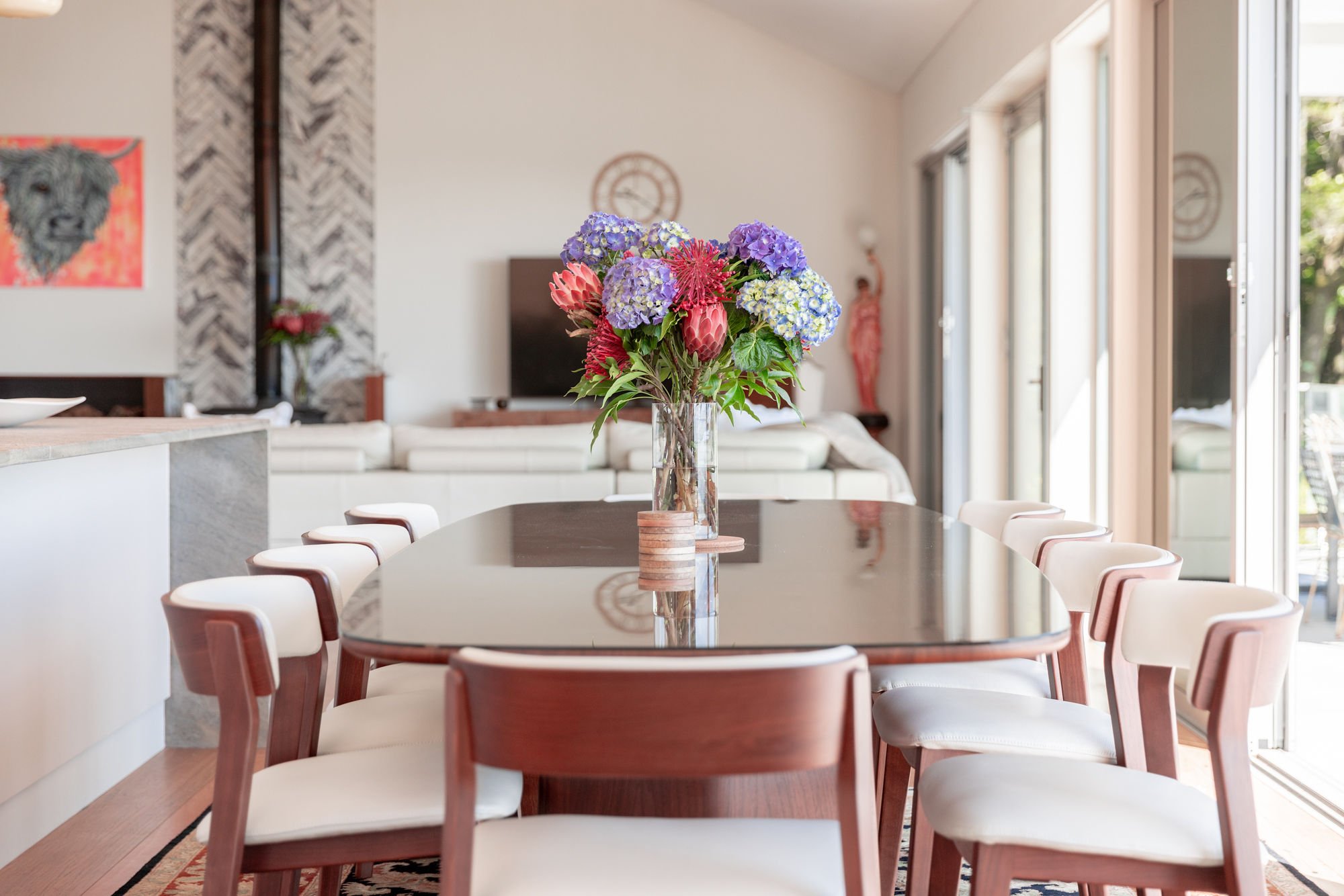Dining area with a large, oval table reflecting light, surrounded by white chairs with wooden backs. A tall clear glass vase filled with colorful flowers, including purple hydrangeas and pink proteas, is on the center of the table. The room has large