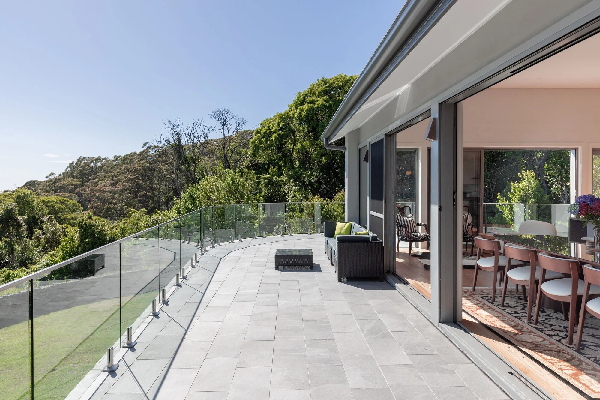 A spacious outdoor balcony with gray tiled flooring and a glass railing, overlooking lush green trees and hills under a clear blue sky. The balcony is connected to an indoor living area through open sliding glass doors, with interior visible furnitur