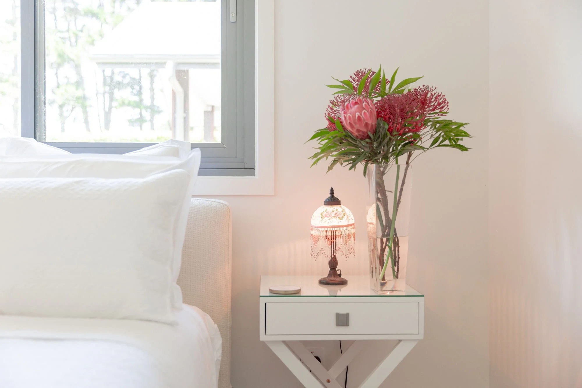 A bedside table with a drawer, holding a glass vase with pink flowers and green leaves, and a decorative table lamp with a floral design, positioned next to a white bed and a window.