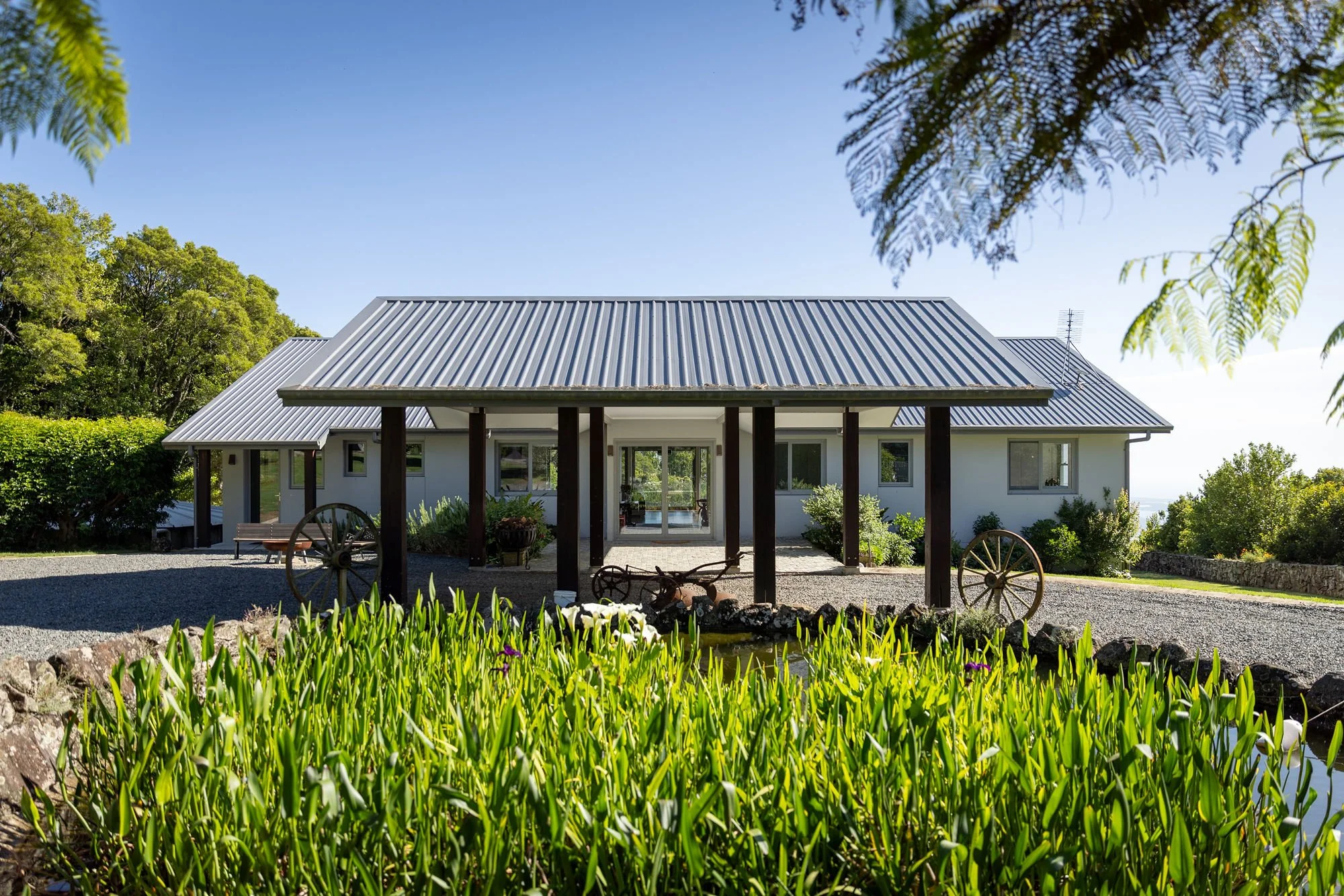 A white house with a metal roof surrounded by greenery, including trees and plants, under a clear blue sky.