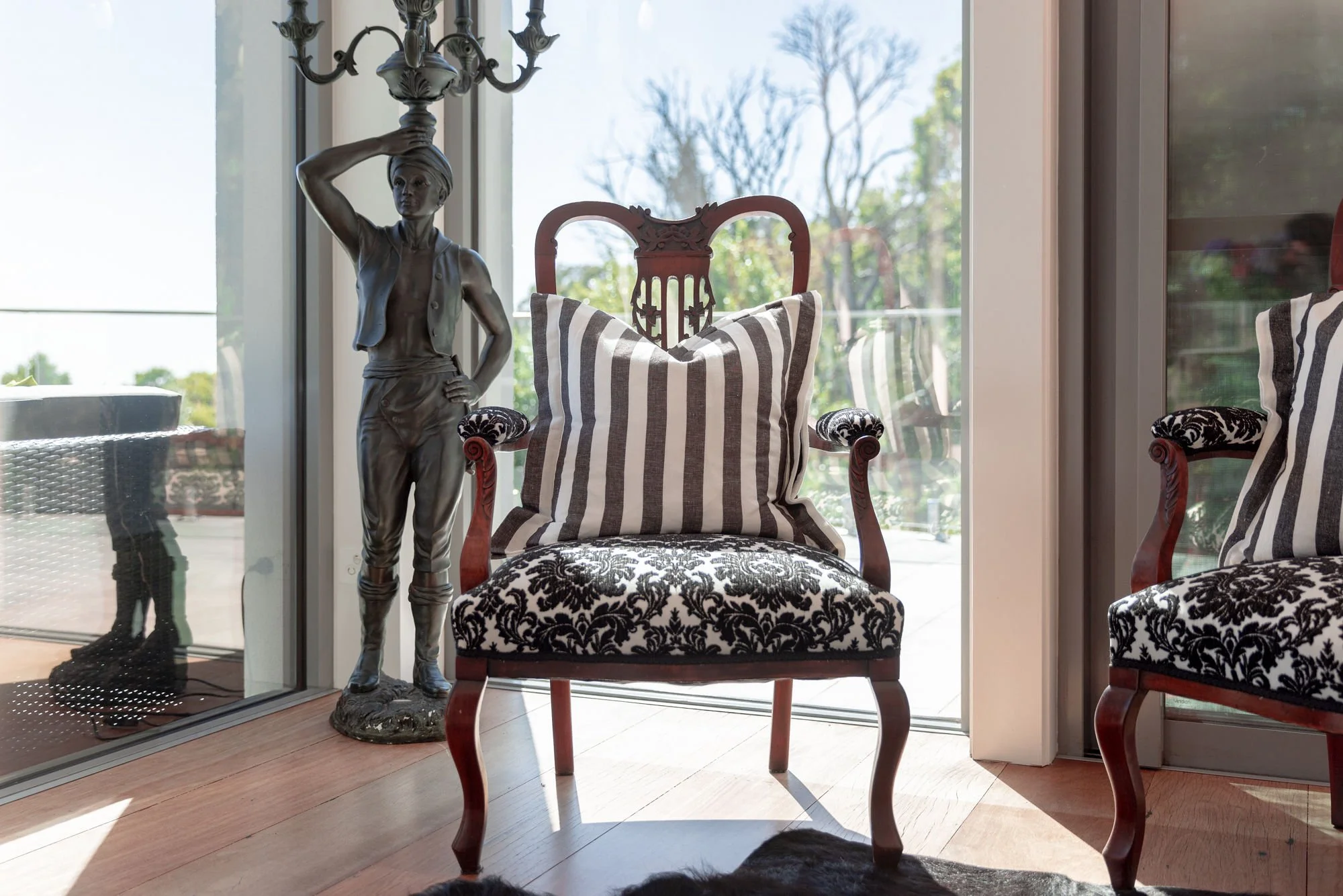A living room scene featuring a vintage wooden chair with a striped pillow, a matching cushion on another chair to the right, a decorative statue of a woman holding a chandelier above her head, and a sliding glass door with a view of trees outside.