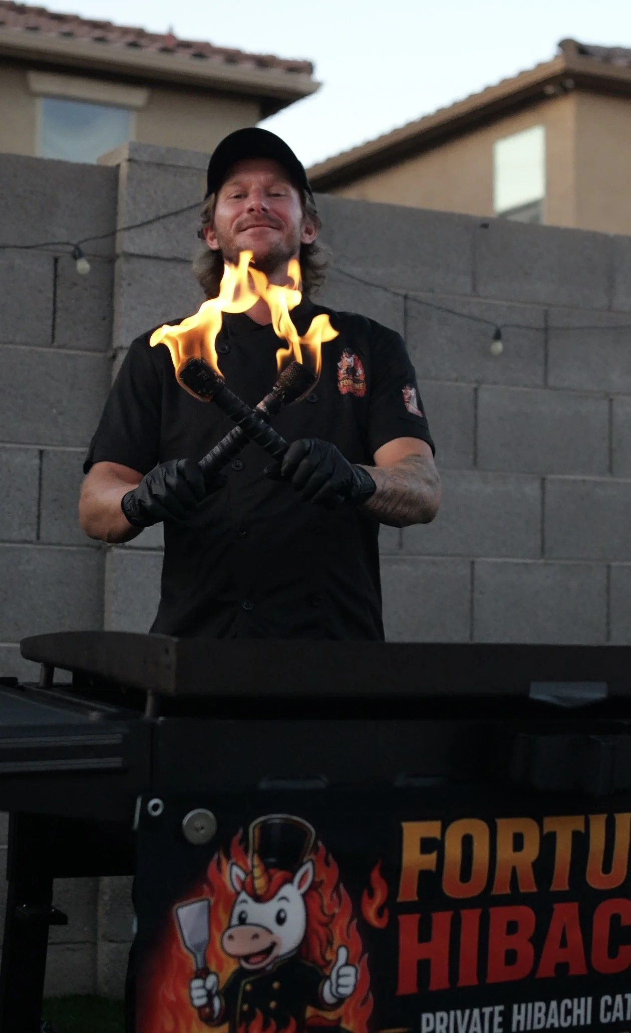 Person holding two flaming torches in front of a privacy booth with a colorful unicorn mascot and the text 'Fortune Hibachi' in an outdoor setting.