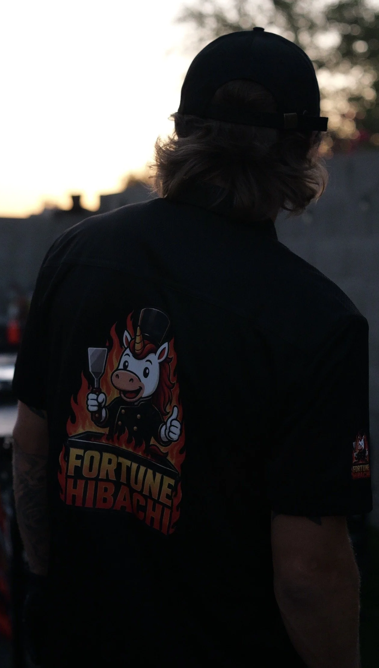 Person wearing a black t-shirt with a cartoon unicorn chef holding a spatula and a frying pan, with a fiery background and the words "Fortune Hibachi" on it, seen from behind during sunset.