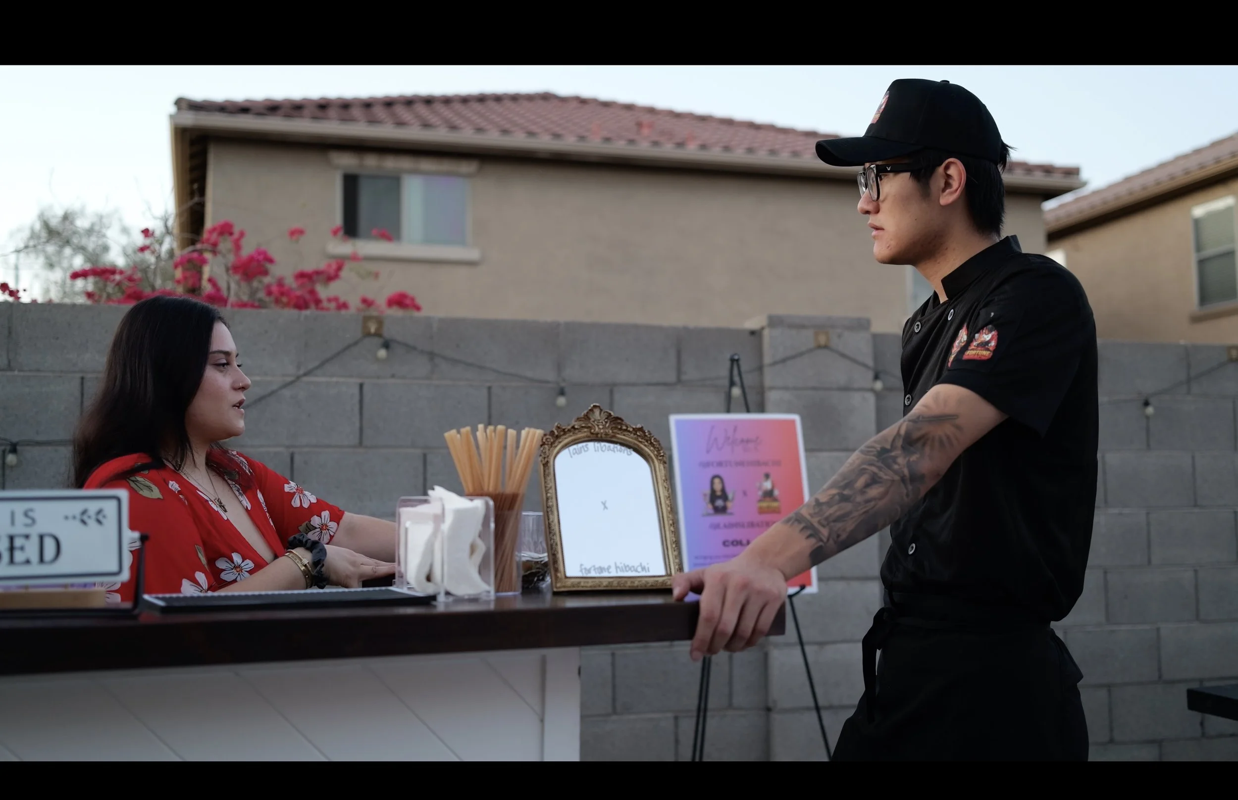 A woman in a red floral dress sitting at a table, talking to a man standing across from her. The man is wearing glasses, a black baseball cap, and a black chef's jacket, with tattoos on his arm. They are outdoors with a gray concrete wall and residential houses in the background.