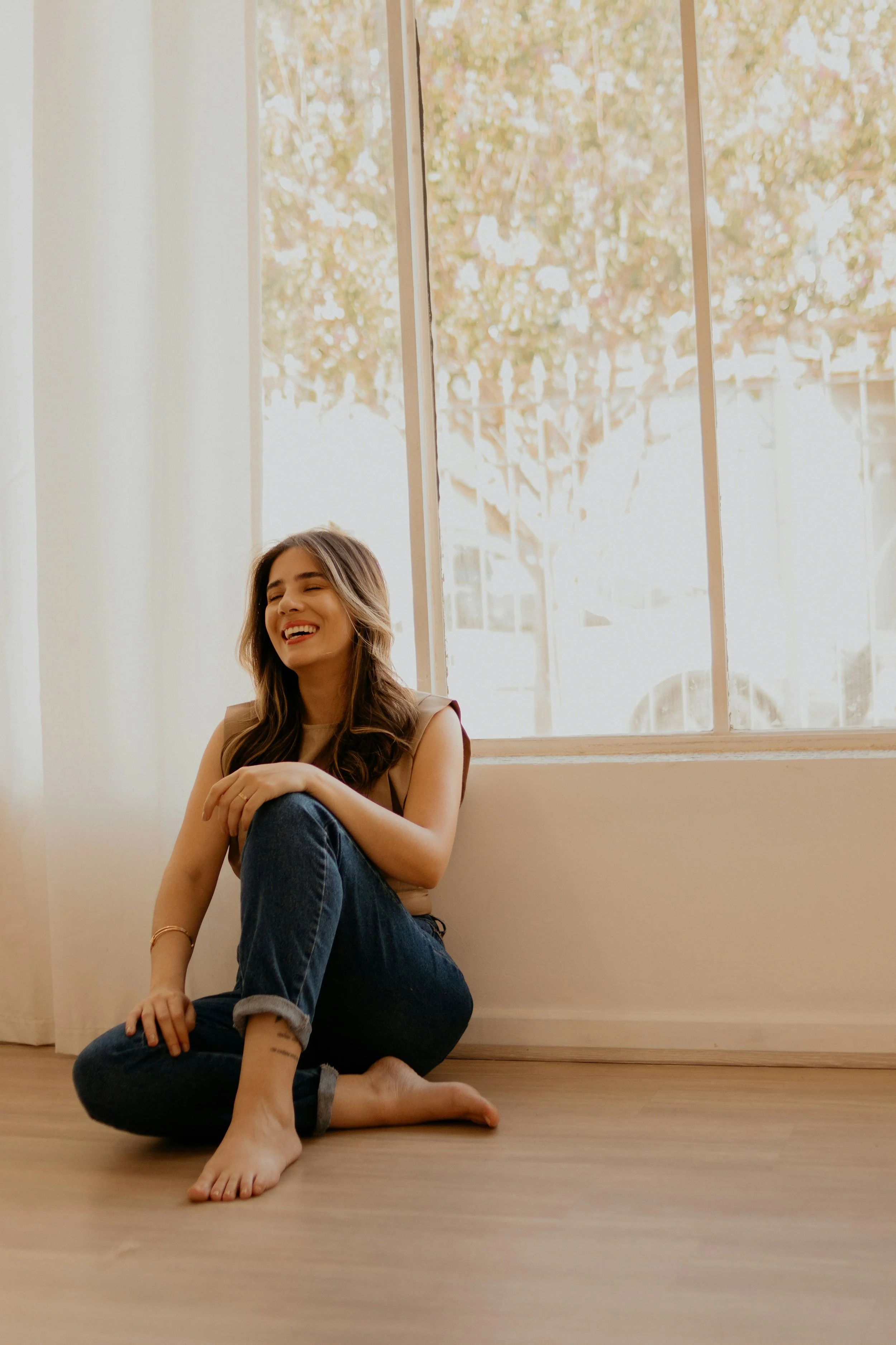 A young woman with long brown hair, sitting on a wooden floor near a large window, smiling and laughing. She is wearing a sleeveless beige top, blue jeans, and has her tattooed ankle visible. The window shows blurred trees outside, and sunlight pours into the room.