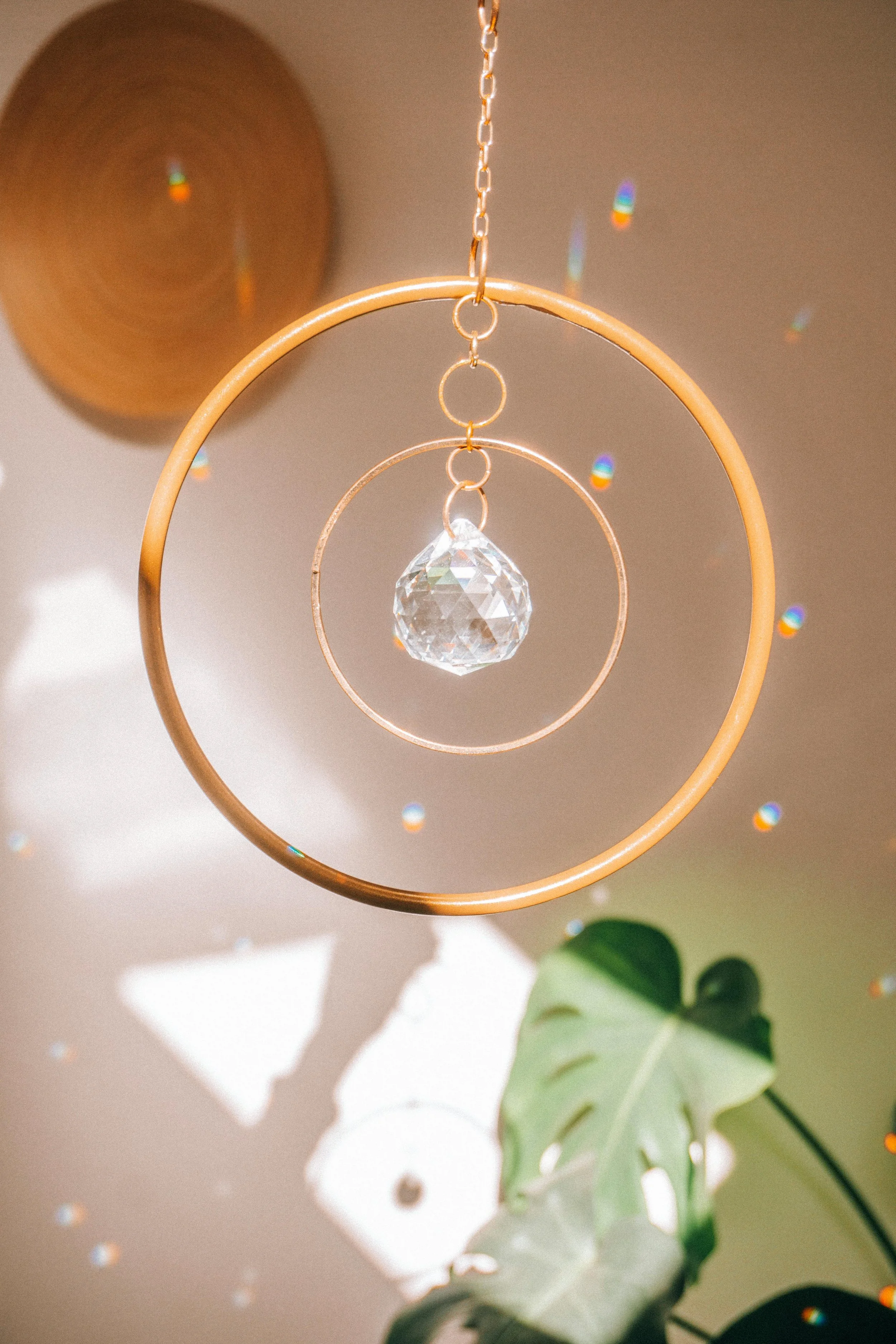A decorative hanging ornament with a clear crystal ball in the center, surrounded by concentric metal rings, suspended from a chain. Behind the ornament, there is a beige ceiling with brightly colored light reflections, and a green monstera leaf is visible in the lower right corner.