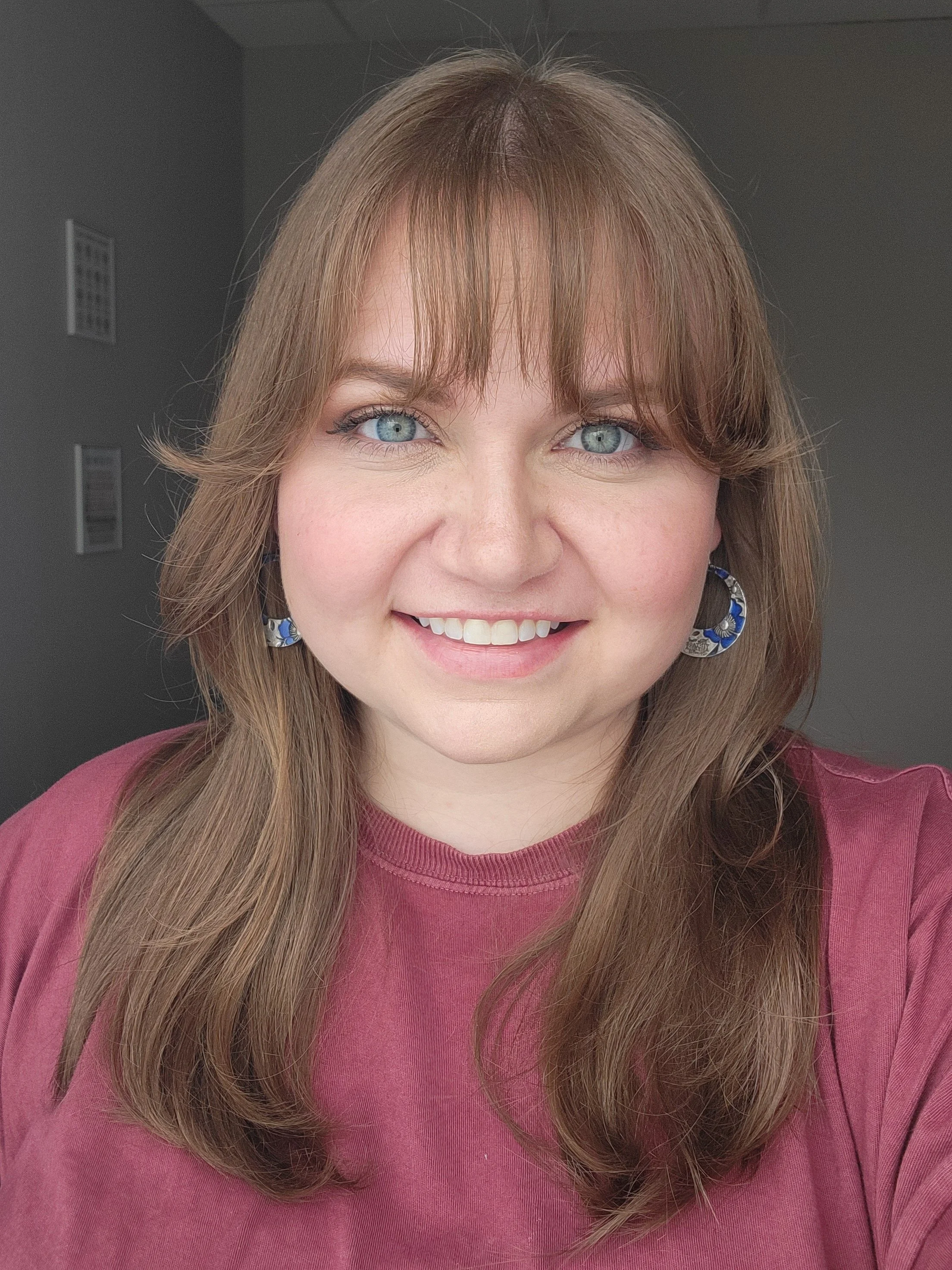 Close-up portrait of a woman with long, wavy reddish-brown hair, wearing large, patterned earrings, and a pinkish-red top, smiling and showing her teeth.