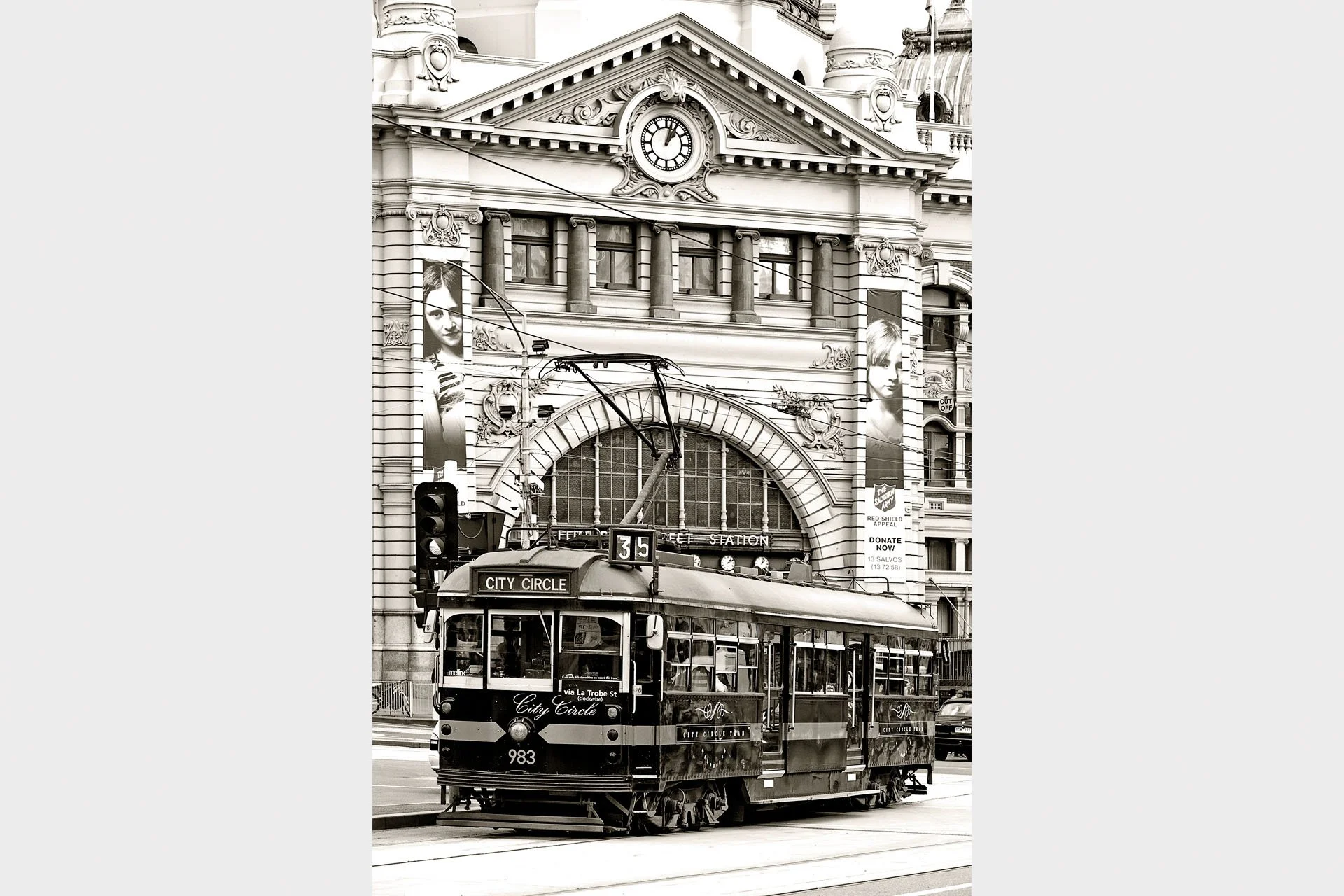Black and white photo of a vintage W-class City Circle tram passing the arched entrance and clock of Flinders Street Station, Melbourne, Victoria.