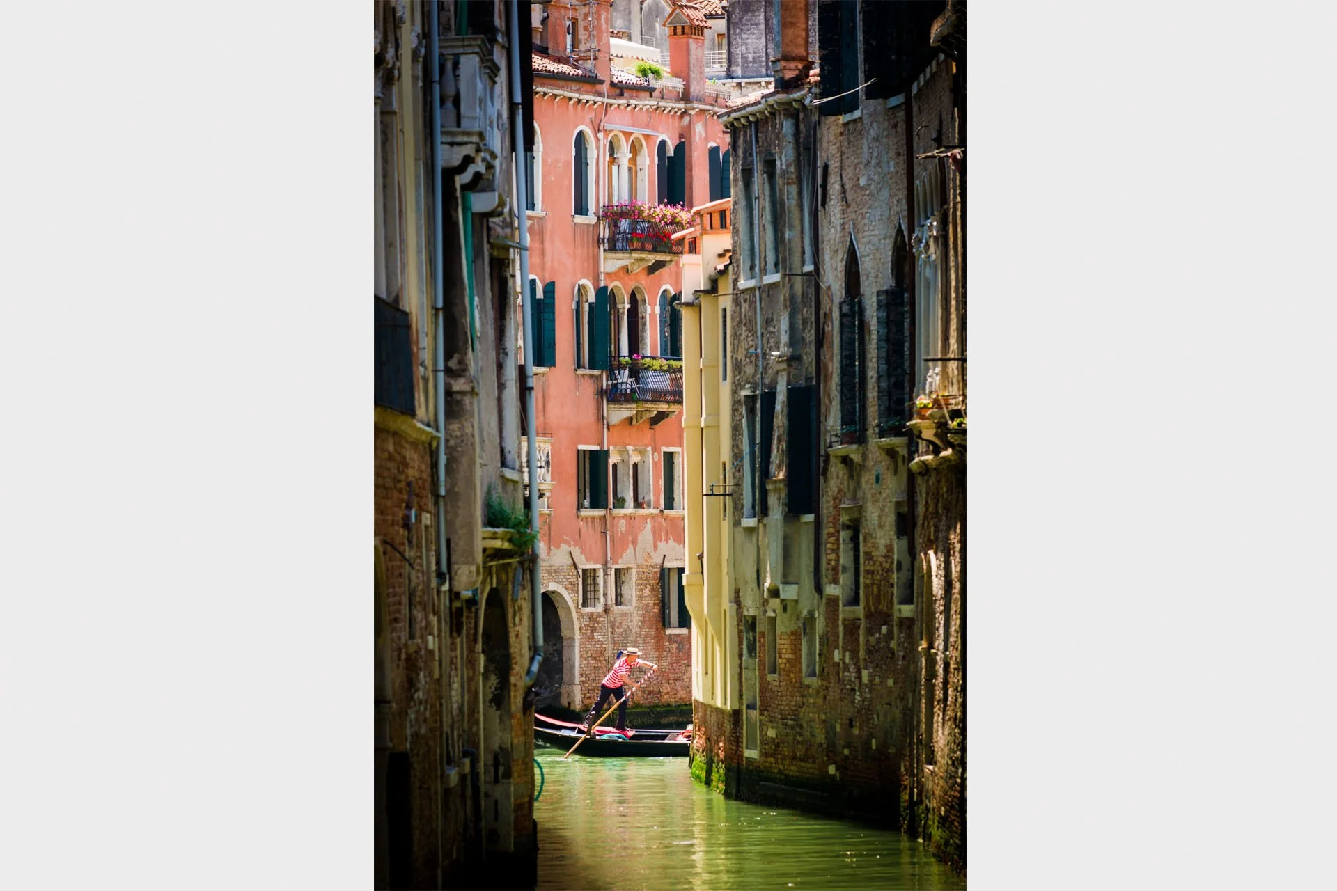 Gondolier in a striped shirt and straw hat rowing a gondola along a narrow green canal between weathered brick and pink palazzos with flower-filled balconies, Venice, Italy.