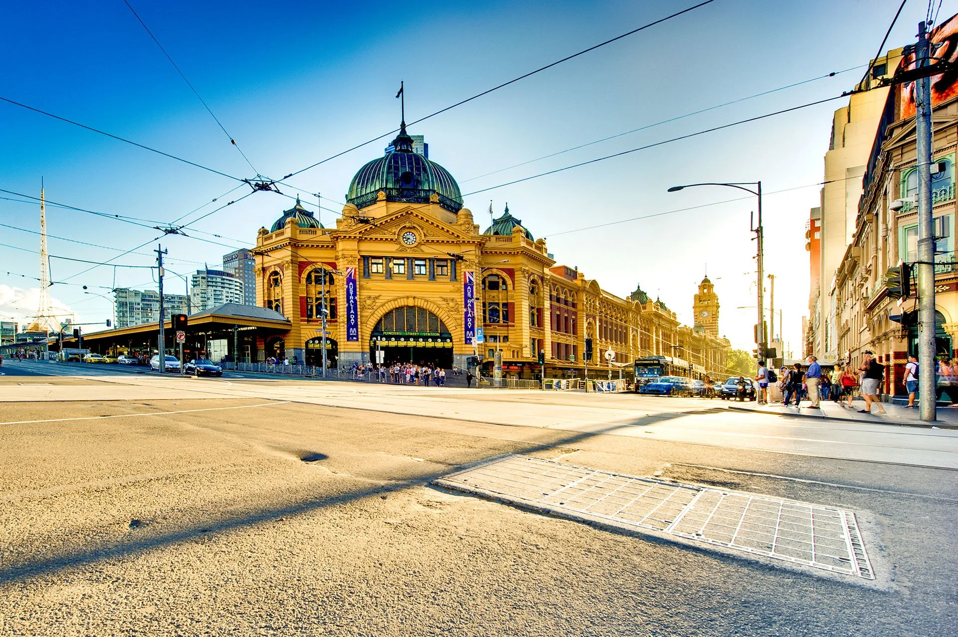 Flinders Street Station in Melbourne, Victoria, glowing gold in late afternoon light, seen from the corner of Flinders and Swanston Streets with tram wires overhead and the Arts Centre spire in the di