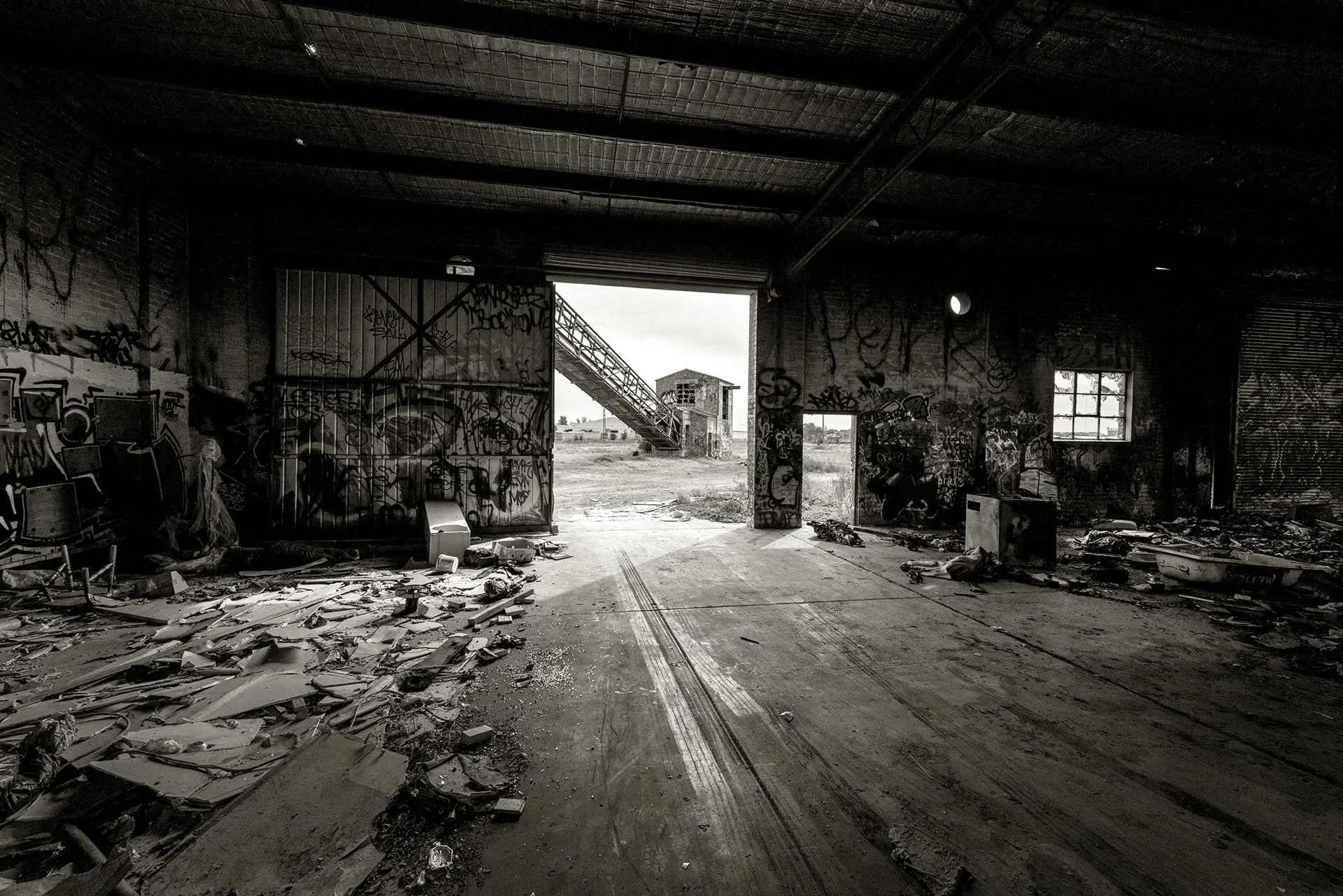 Black and white photo inside the abandoned Bradmill textile factory in Yarraville, Melbourne, with graffiti-covered walls, debris-strewn floor and a large doorway opening onto derelict outbuildings.