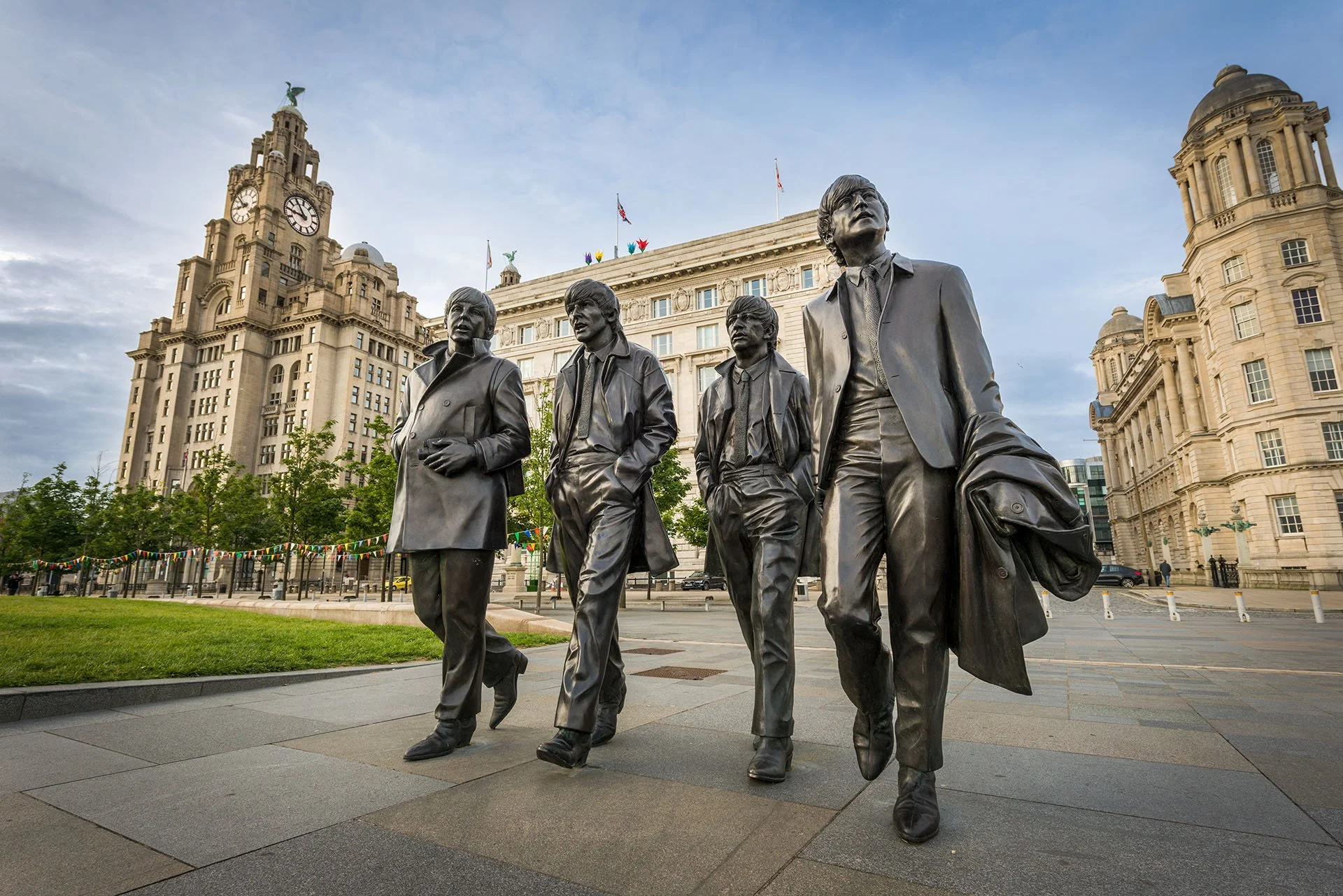 Bronze statue of the Beatles striding across Pier Head in Liverpool, England, with the Royal Liver Building and Cunard Building behind under a soft blue sky.