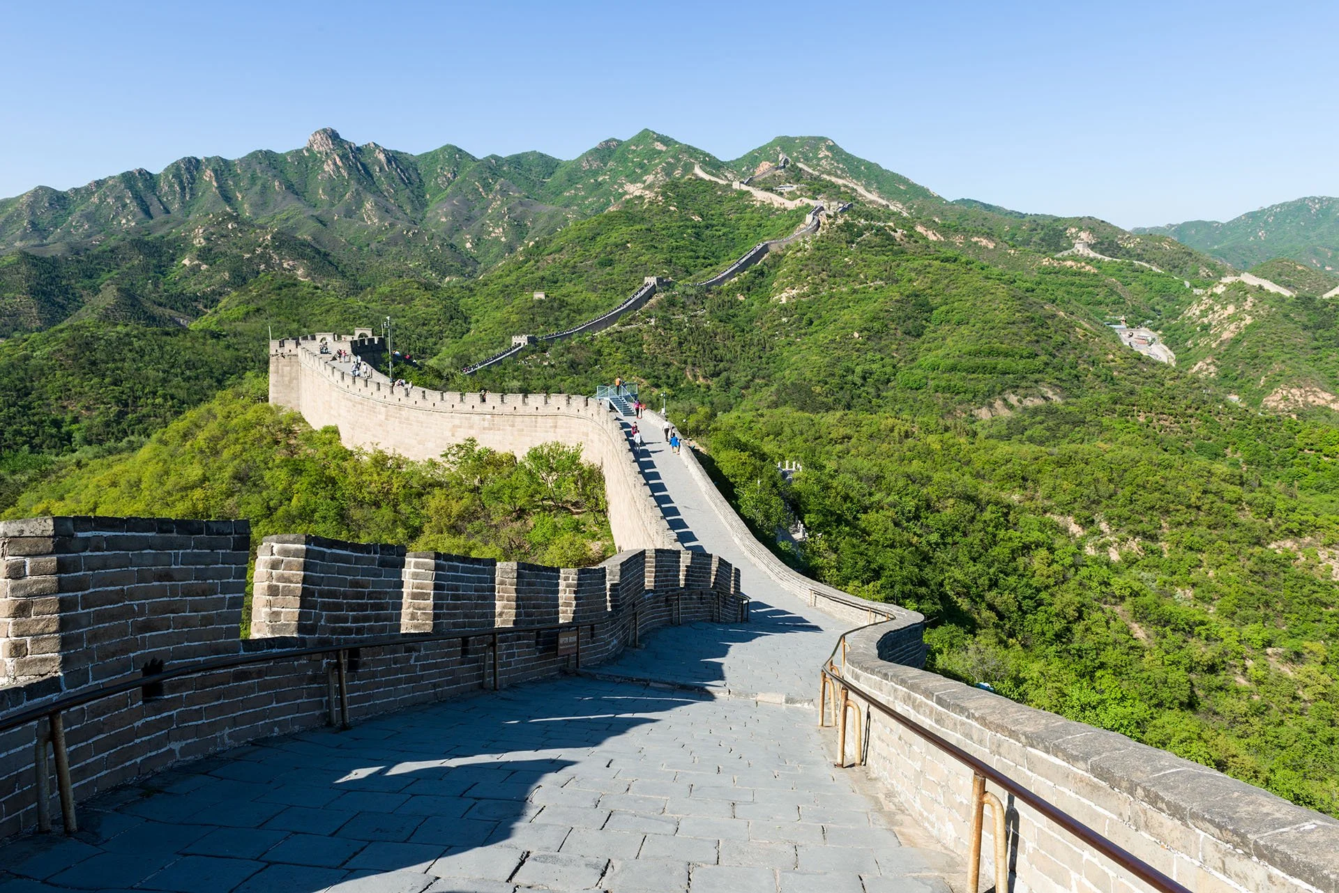 The Great Wall of China winding along green mountain ridges at Badaling, near Beijing, with its stone walkway and battlements stretching into the distance under a clear blue sky.