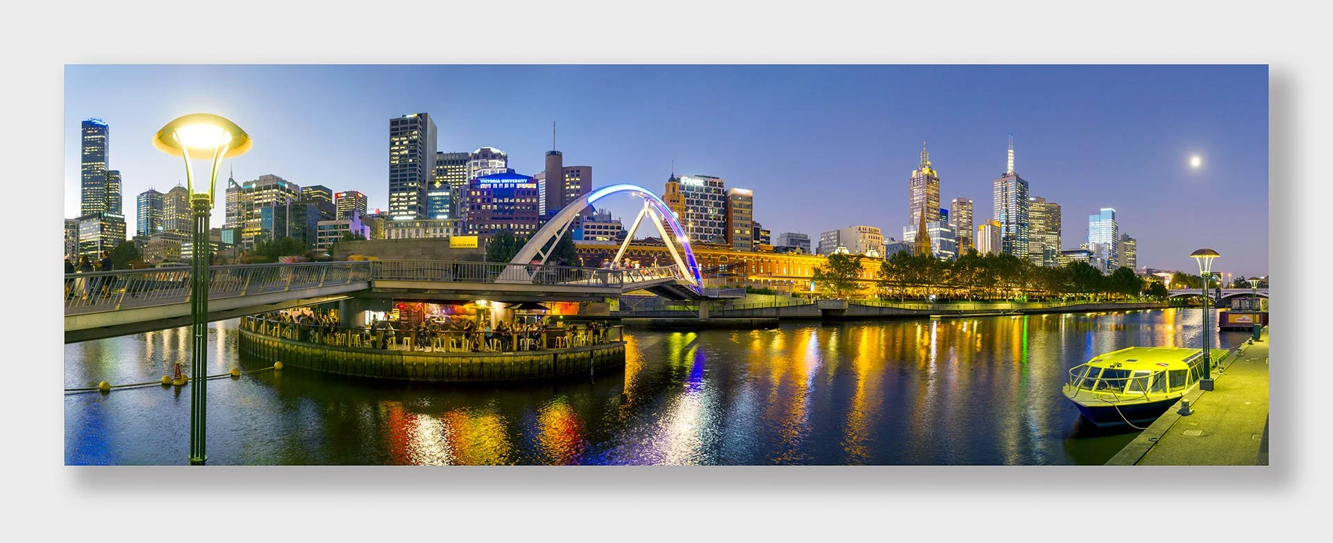 Panorama of the Yarra River at dusk, Melbourne, Victoria, with the lit Evan Walker Bridge, Arbory Afloat bar, Flinders Street Station and city towers reflected in the water.