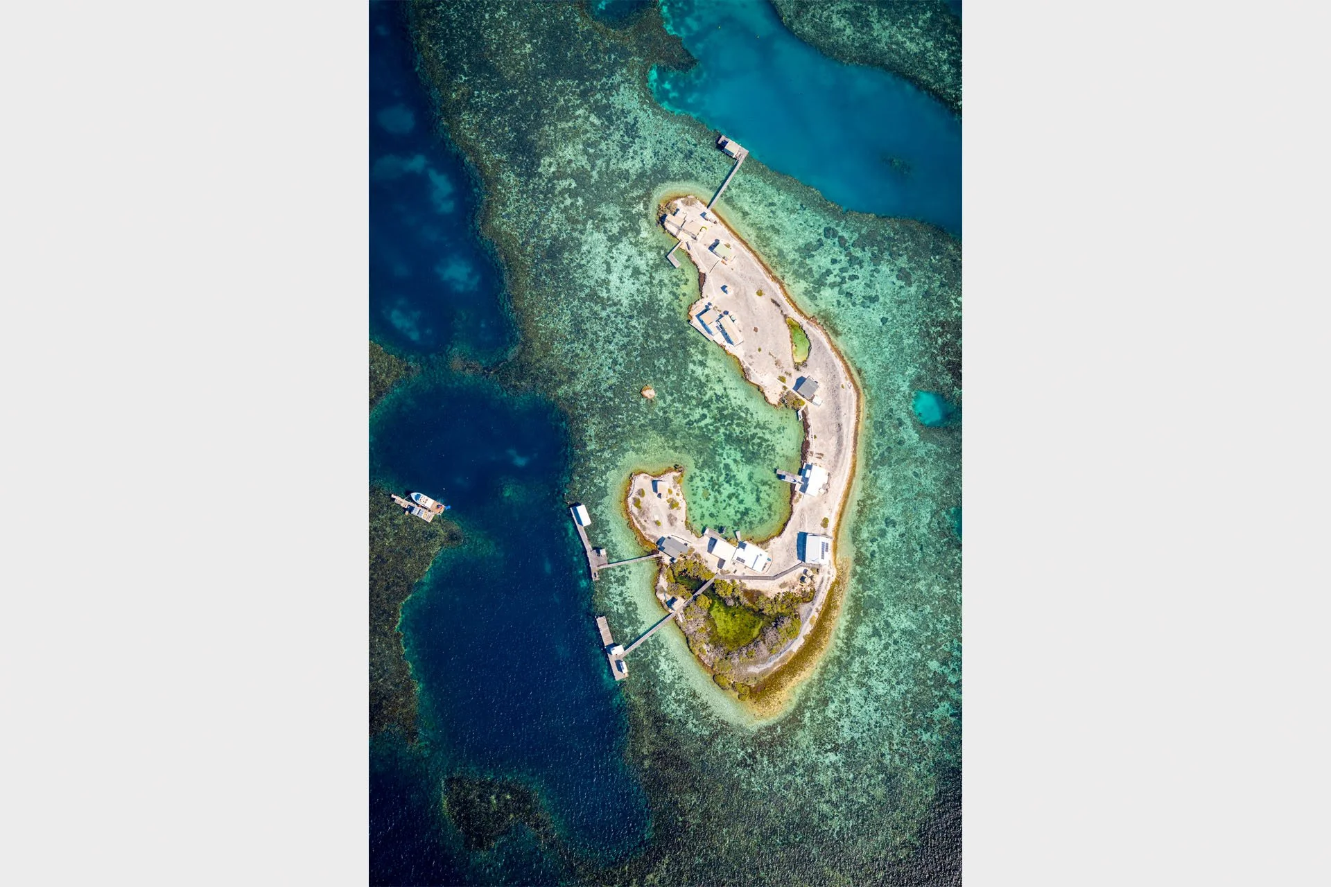 Aerial view looking down on crescent-shaped Burnett Island in the Houtman Abrolhos Islands, Western Australia, with fishing huts and jetties surrounded by coral reef and deep blue water.