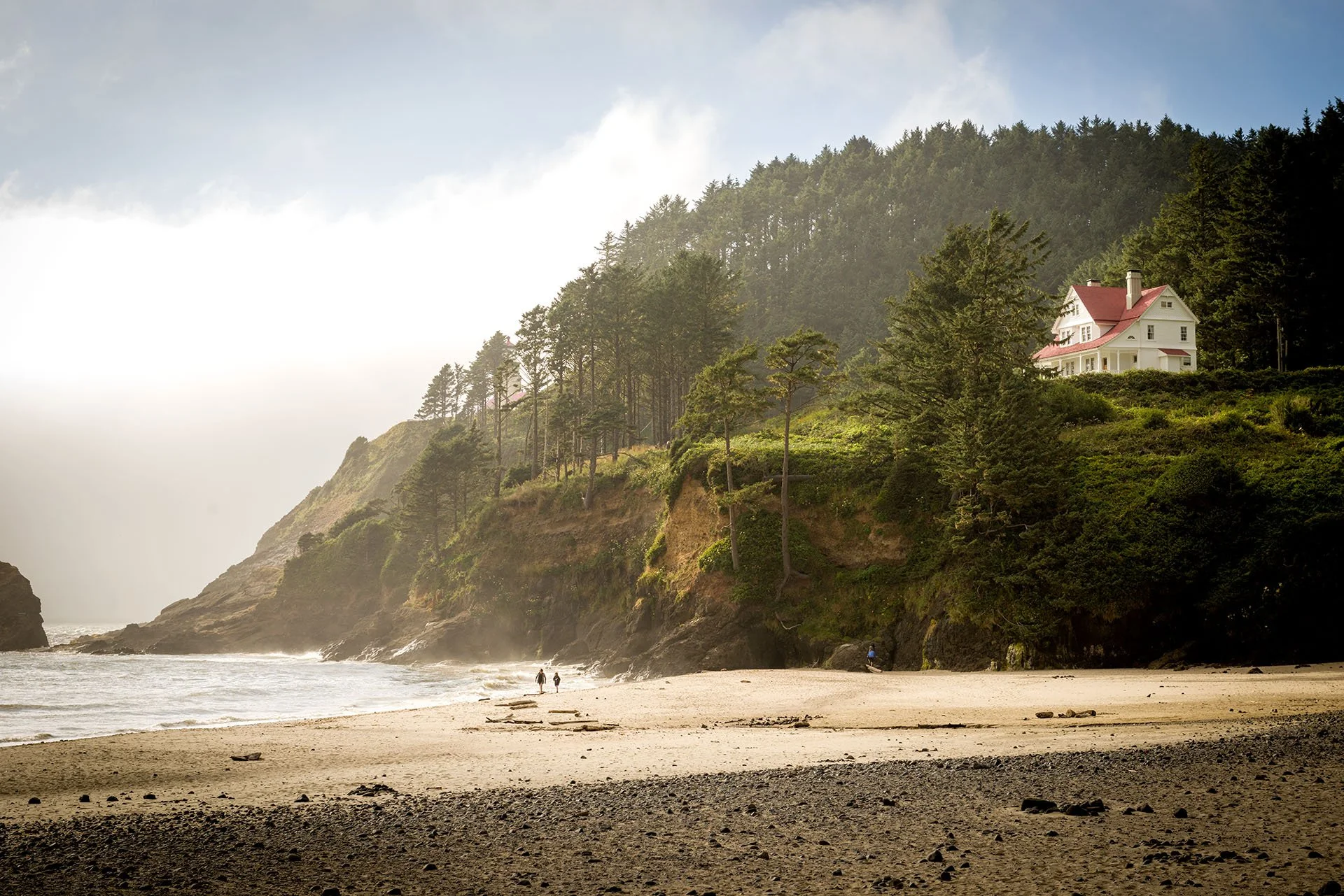 The white Heceta Head lighthouse keeper’s house on a forested headland above a misty beach near Florence on the Oregon coast, USA, with two people walking along the shore.