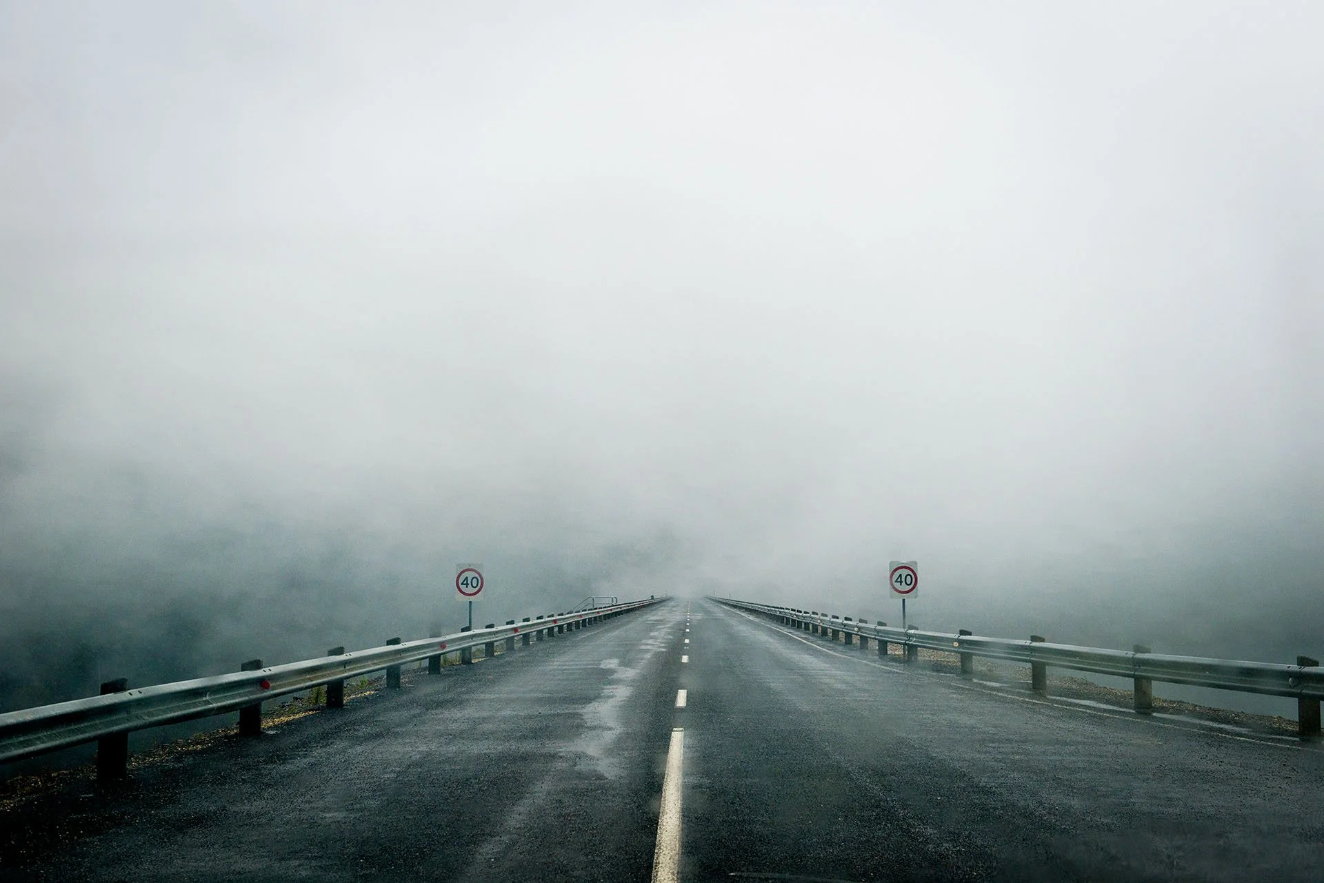 Wet road with guard rails and 40 km/h signs disappearing into thick fog across the dam wall at Lake Eildon, Victoria.