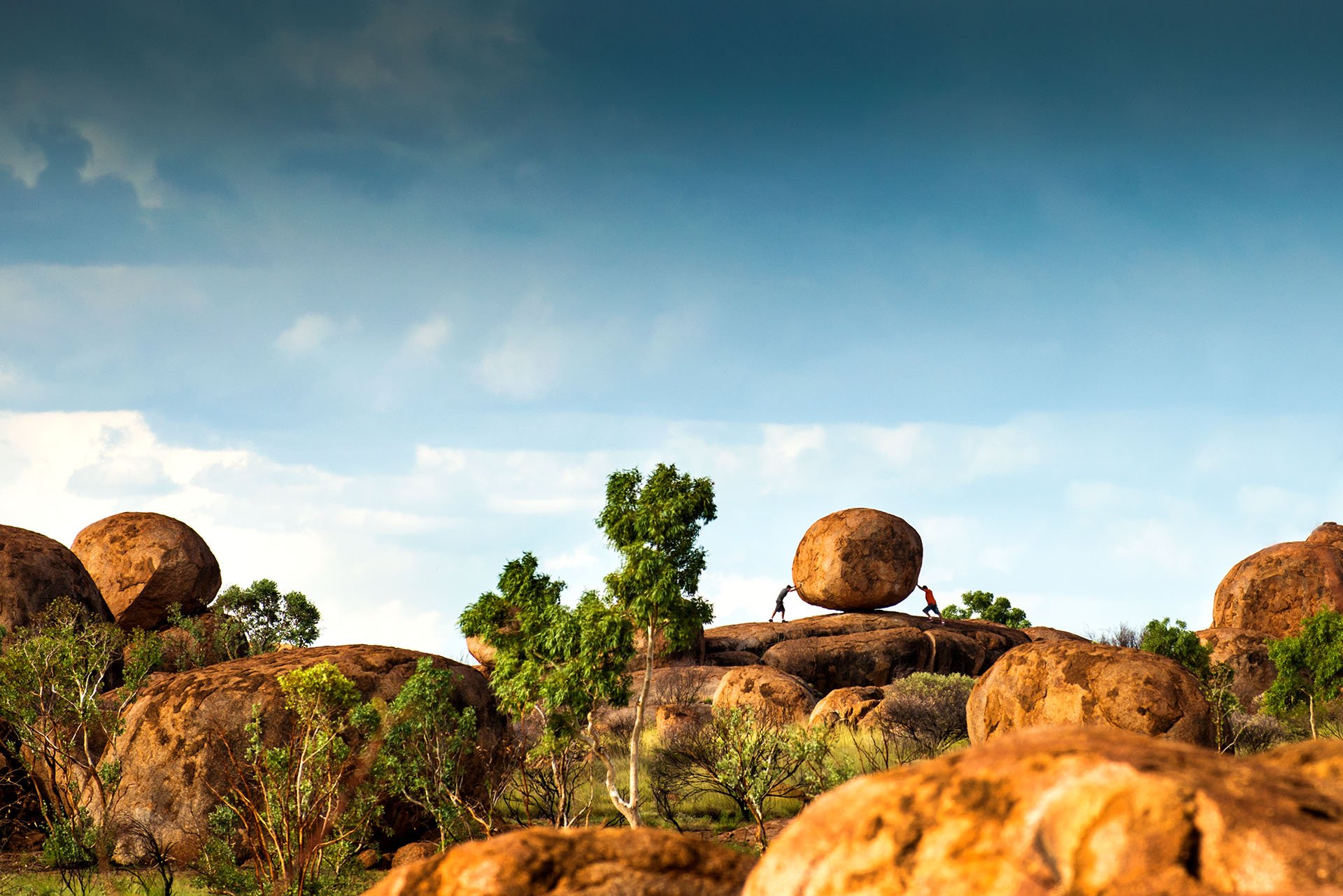 Two people appear to push a giant round granite boulder from opposite sides at Karlu Karlu / Devils Marbles Conservation Reserve, Northern Territory, under a stormy blue sky.