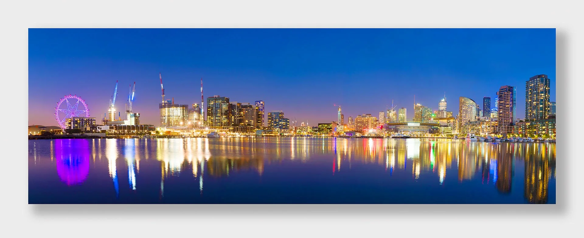 Panorama of Docklands, Melbourne, Victoria, at blue hour, with the Melbourne Star lit purple, city towers, cranes and Marvel Stadium reflected in the still waters of Victoria Harbour.