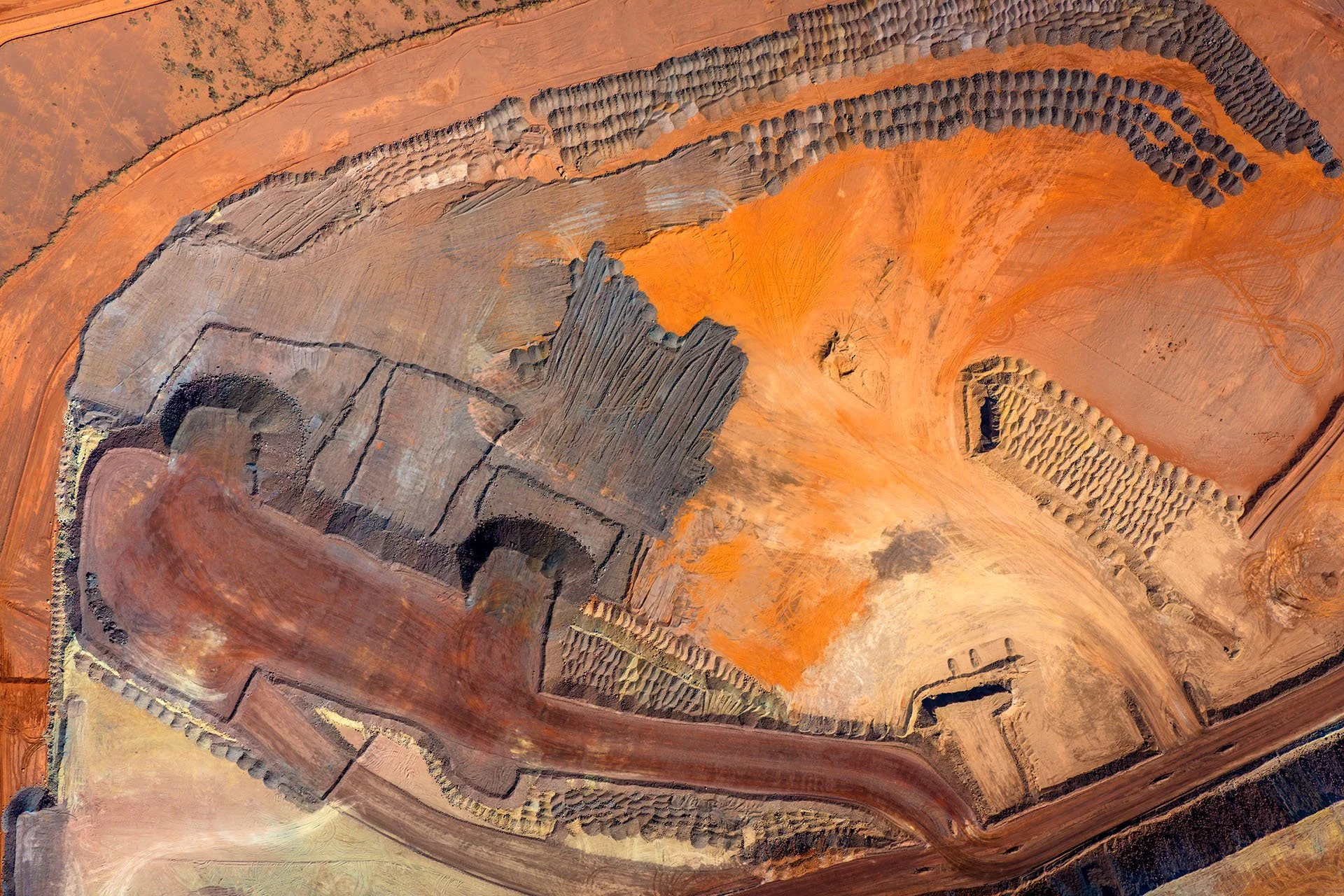 Abstract aerial view of an outback mine site in South Australia, with haul roads, rows of tipped spoil heaps and bands of orange, rust and grey earth.