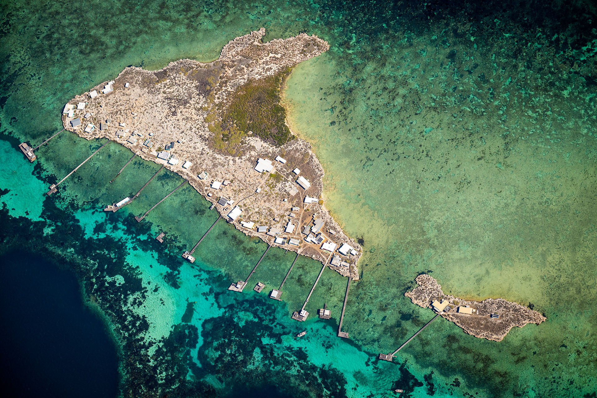 Aerial view of Little Rat and Bushy Joe Islands in the Houtman Abrolhos Islands, Western Australia, with fishing huts and long jetties reaching into turquoise reef waters.