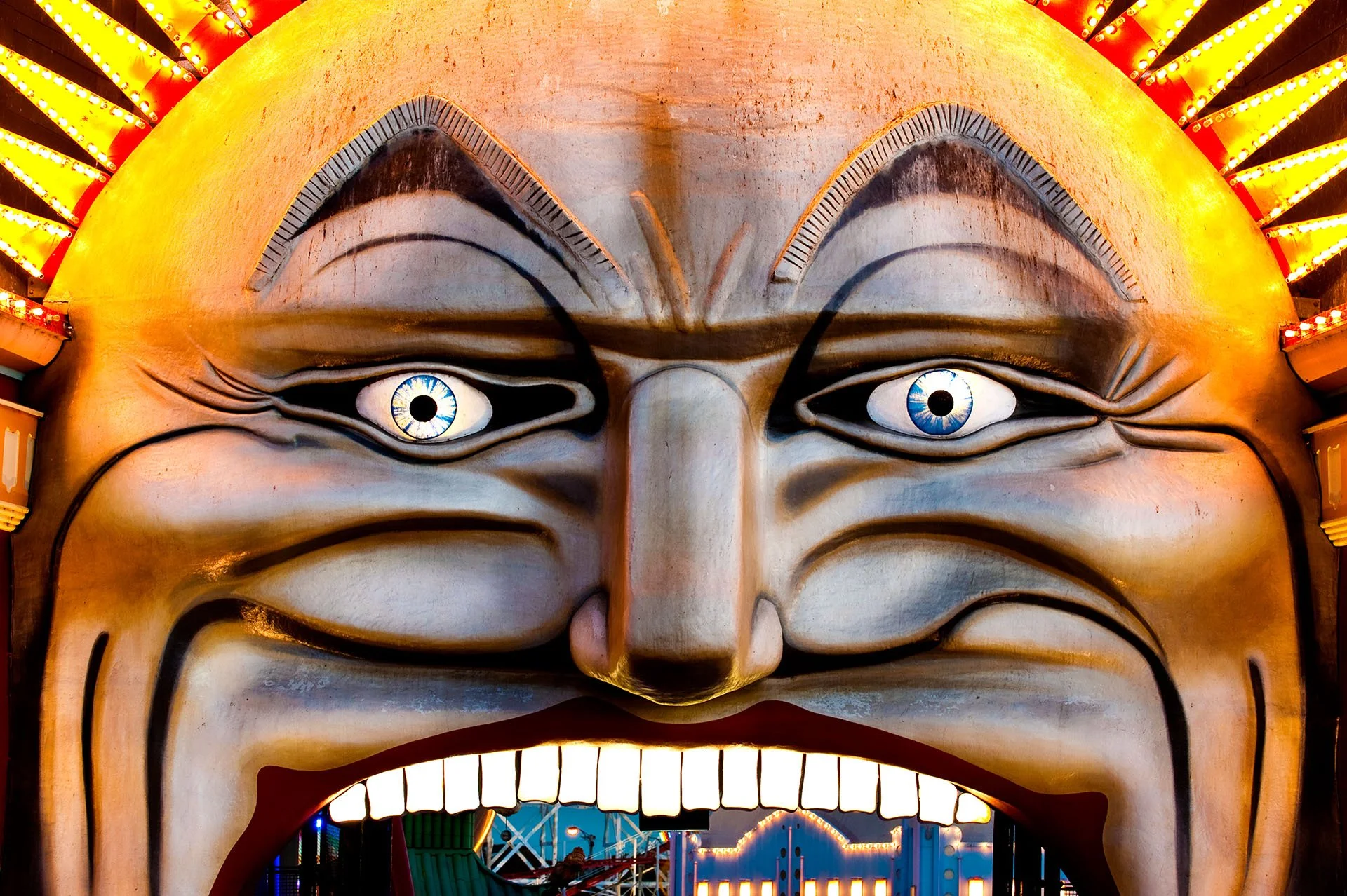 Close-up of the giant grinning face entrance to Luna Park, St Kilda, Melbourne, with wide blue eyes, bared teeth and a glowing ring of lights, rides visible through the open mouth.