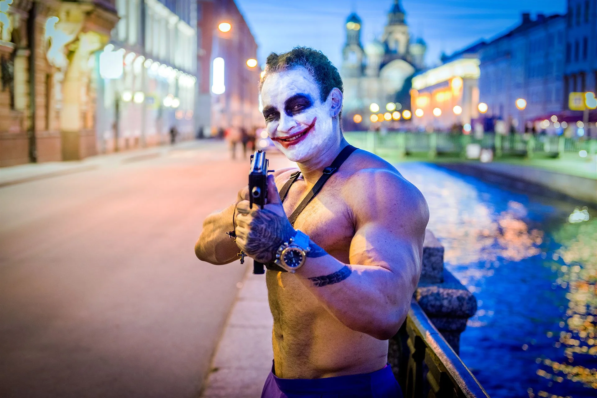 Muscular man in Joker-style face paint aiming a pistol at the camera beside the Griboyedov Canal, St Petersburg, Russia, with the Church of the Saviour on Spilled Blood behind.