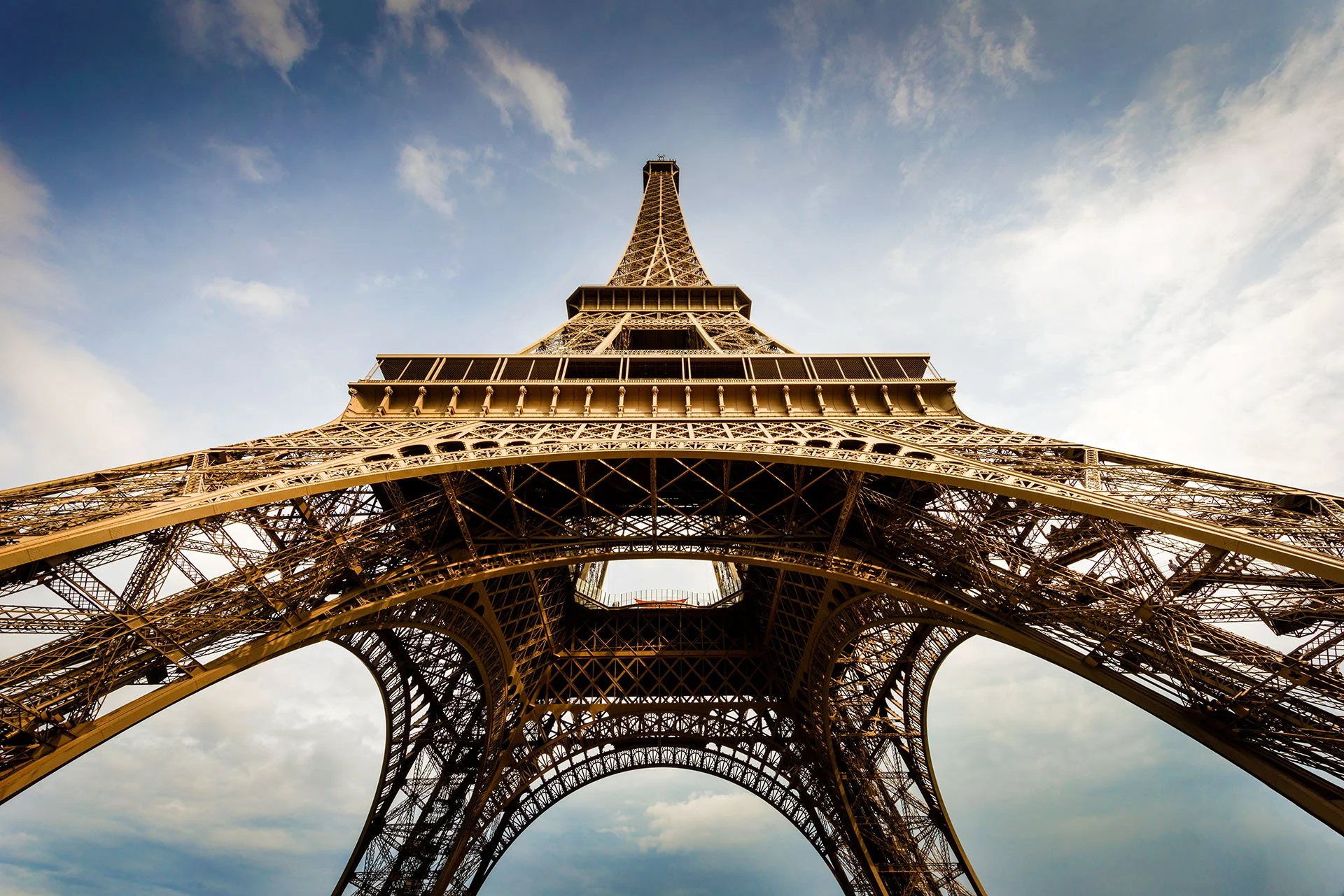 Dramatic wide-angle view looking straight up from beneath the Eiffel Tower in Paris, France, its iron lattice arches framing a blue sky with wispy clouds.