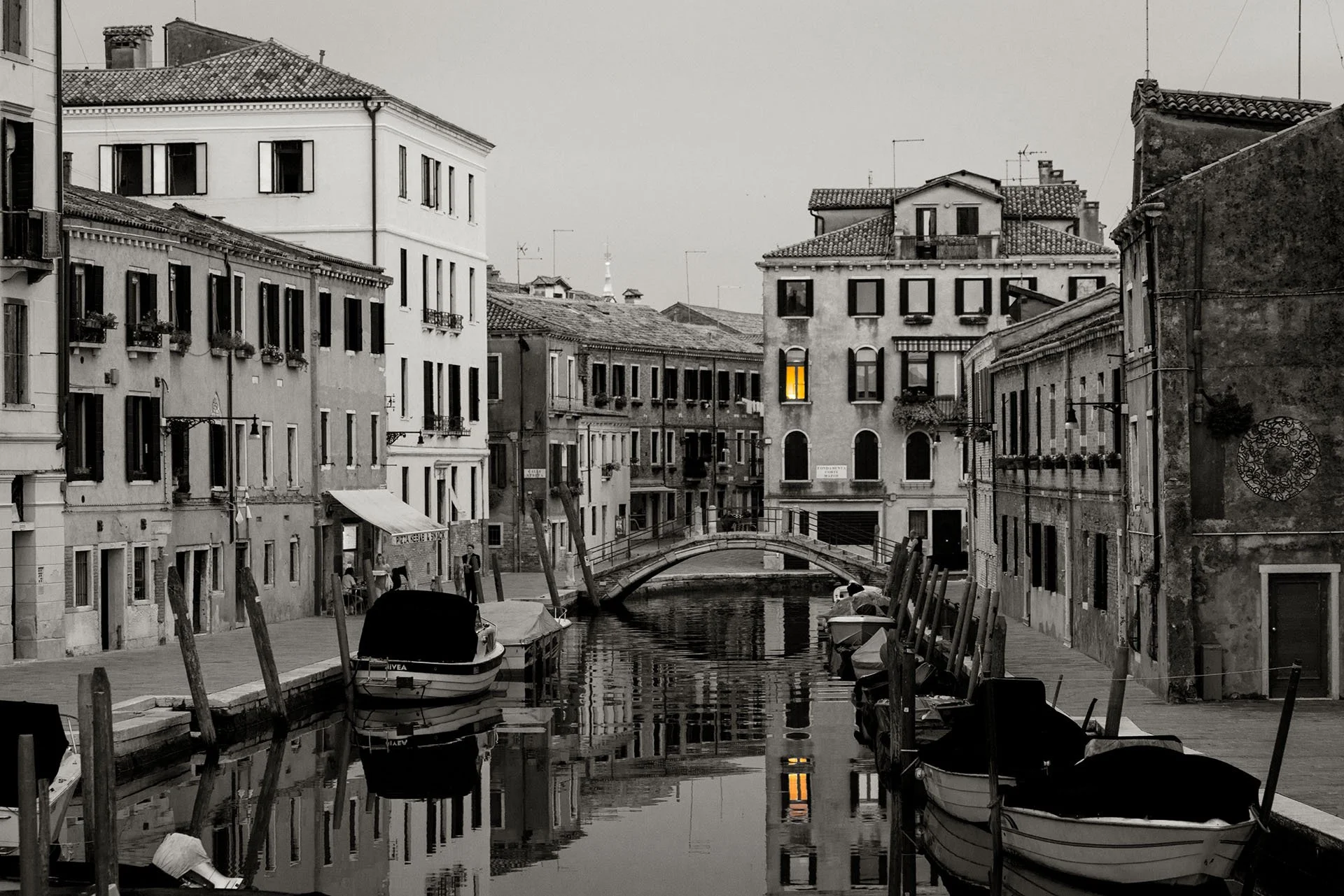 Black and white view along a quiet Venice canal in Italy, with moored boats, a small arched bridge and a single glowing yellow window reflected in the still water.