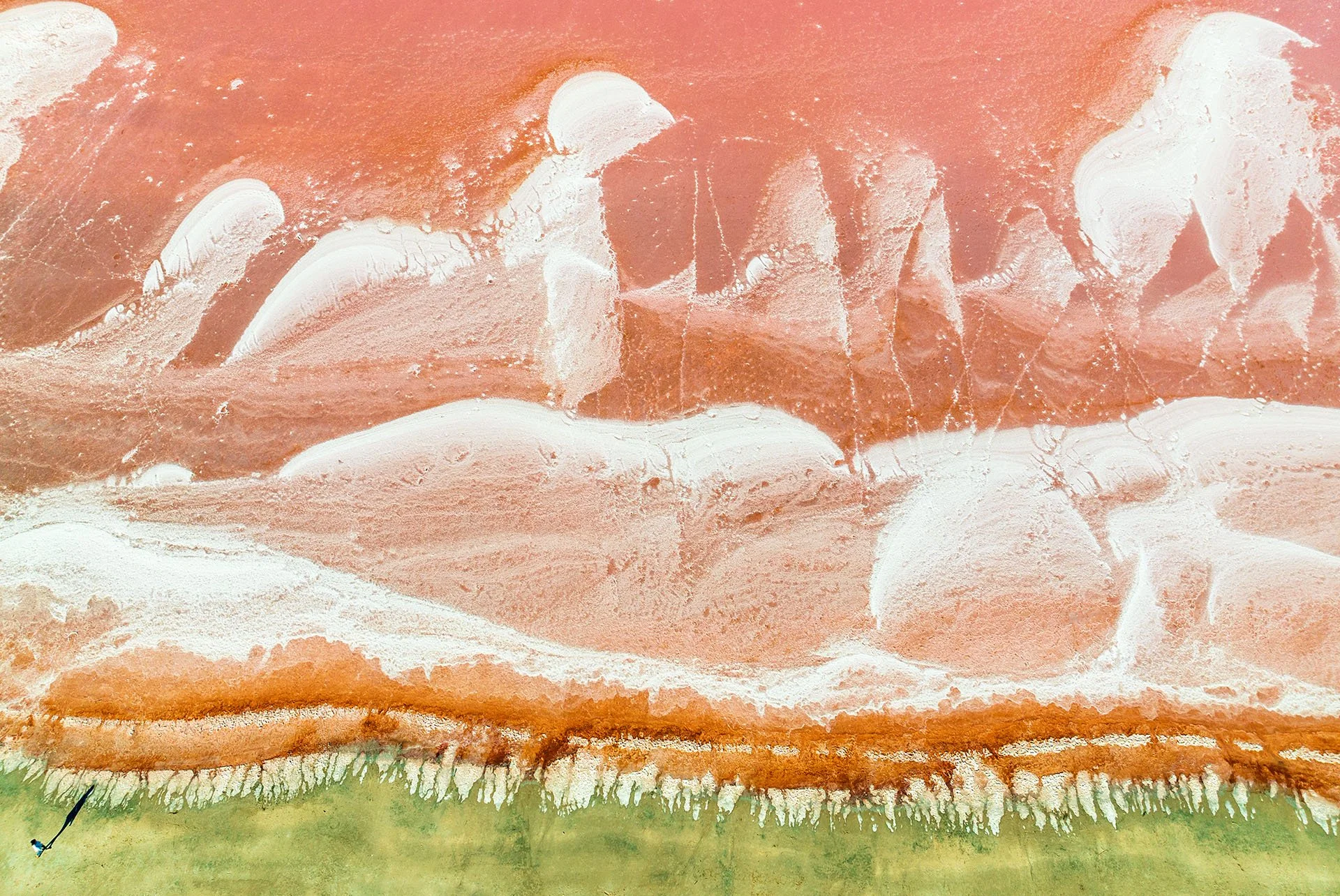 Drone view looking straight down on swirling white salt crust and pink water at the Pink Lake near Dimboola, Victoria, with a tiny figure and long shadow on the green shoreline for scale.