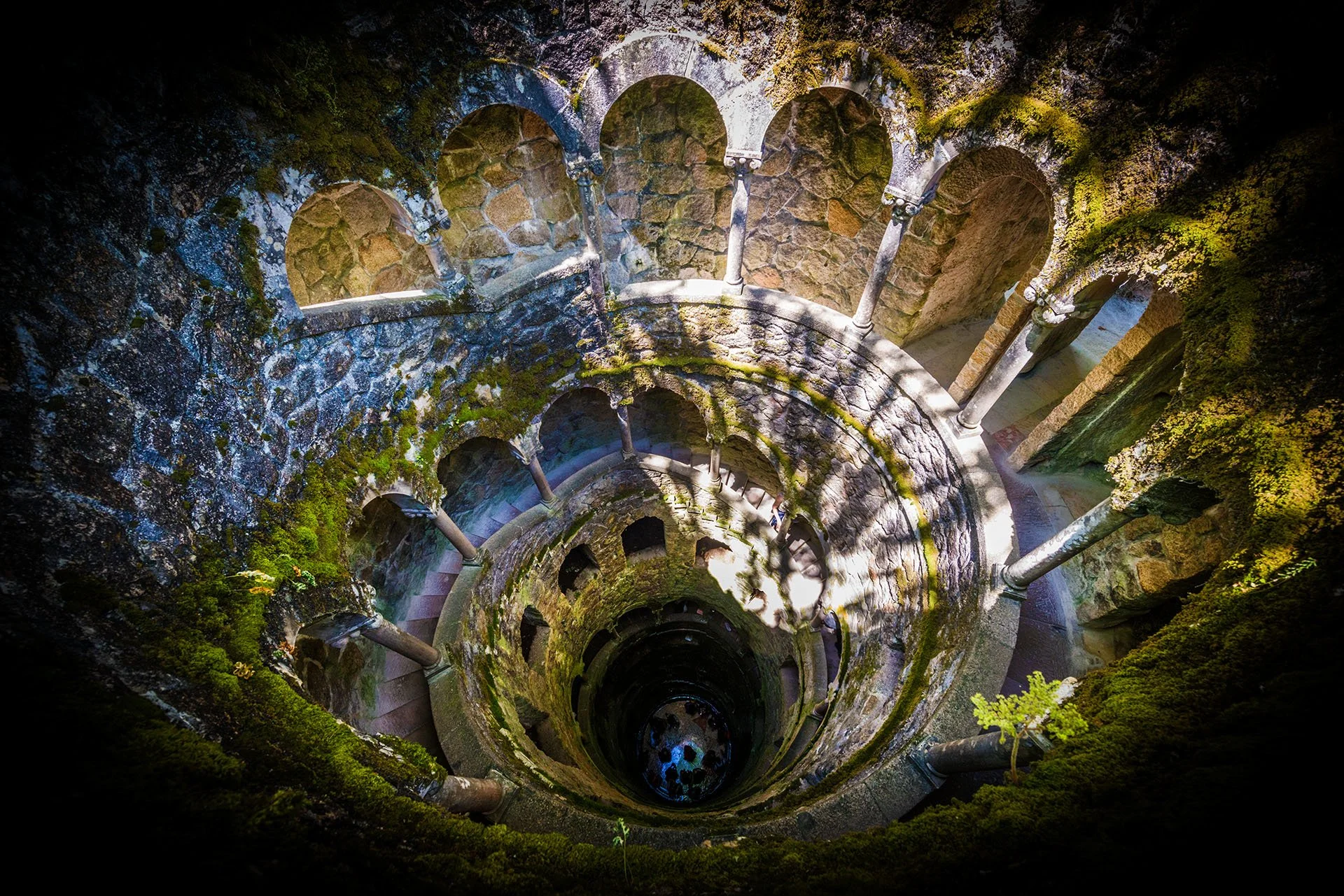 Looking down the spiral staircase of the Initiation Well at Quinta da Regaleira, Sintra, Portugal, with moss-covered stone arches and columns descending into darkness.