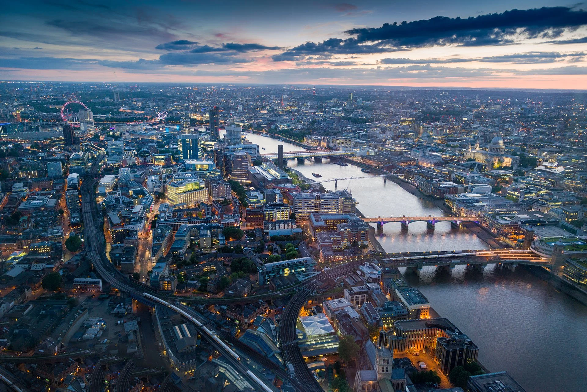 London at dusk from the Shard, looking west up the River Thames past its bridges to St Paul’s Cathedral and the London Eye, with city lights glowing under a pastel sky.