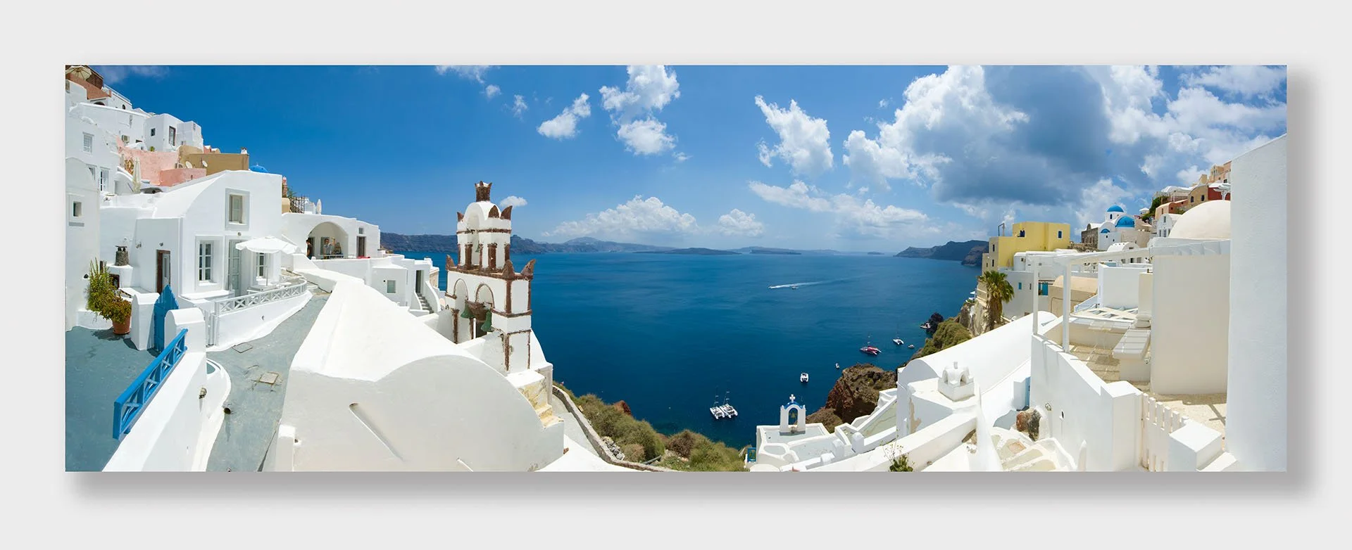 Panorama of whitewashed houses, a bell tower and blue domes in Oia, Santorini, Greece, overlooking the deep blue caldera with boats moored below and white clouds overhead.