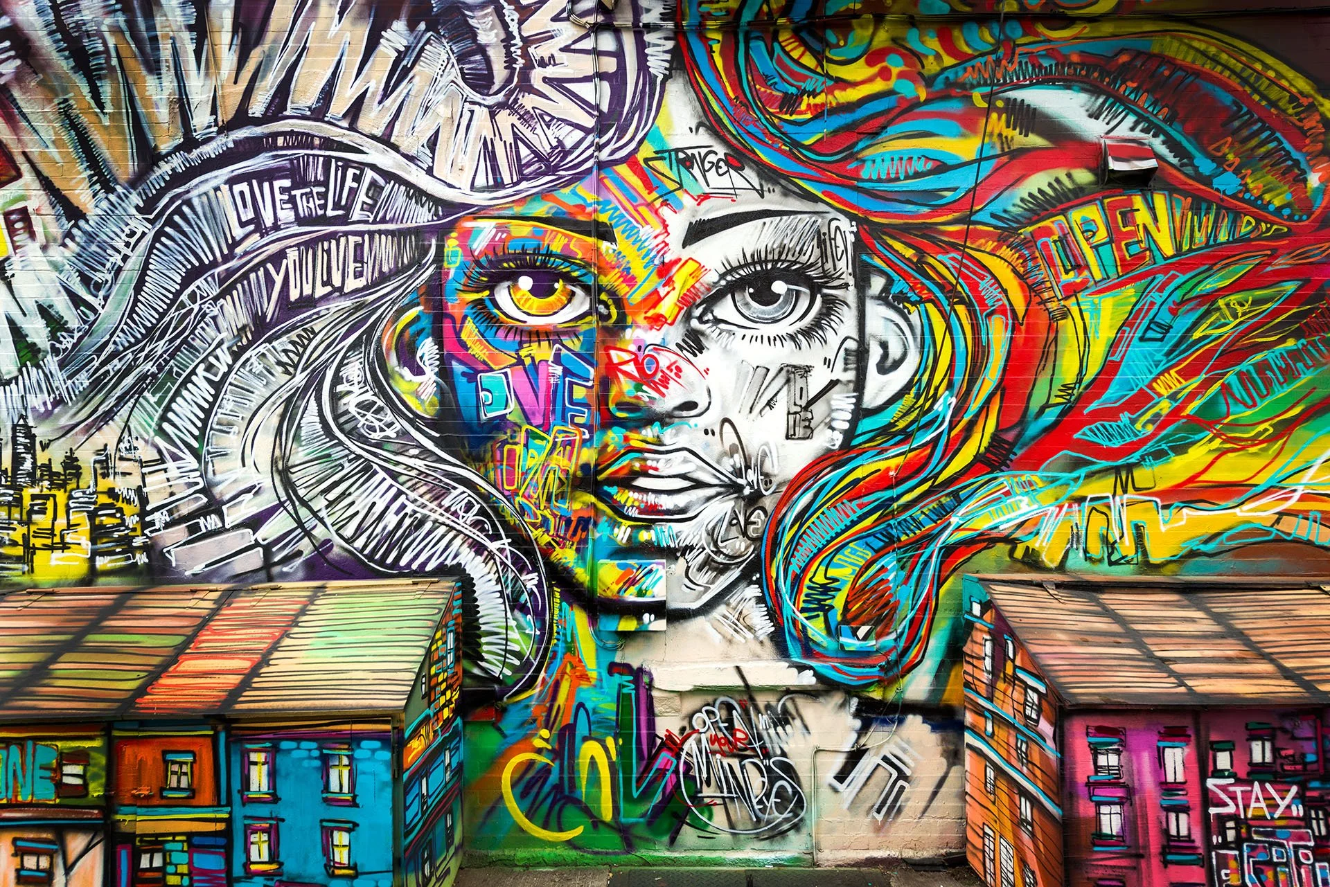 Vibrant street art mural of a woman’s face with flowing rainbow-coloured hair and the words “Love the life you live”, painted above two small shed-like buildings in Brooklyn, New York.