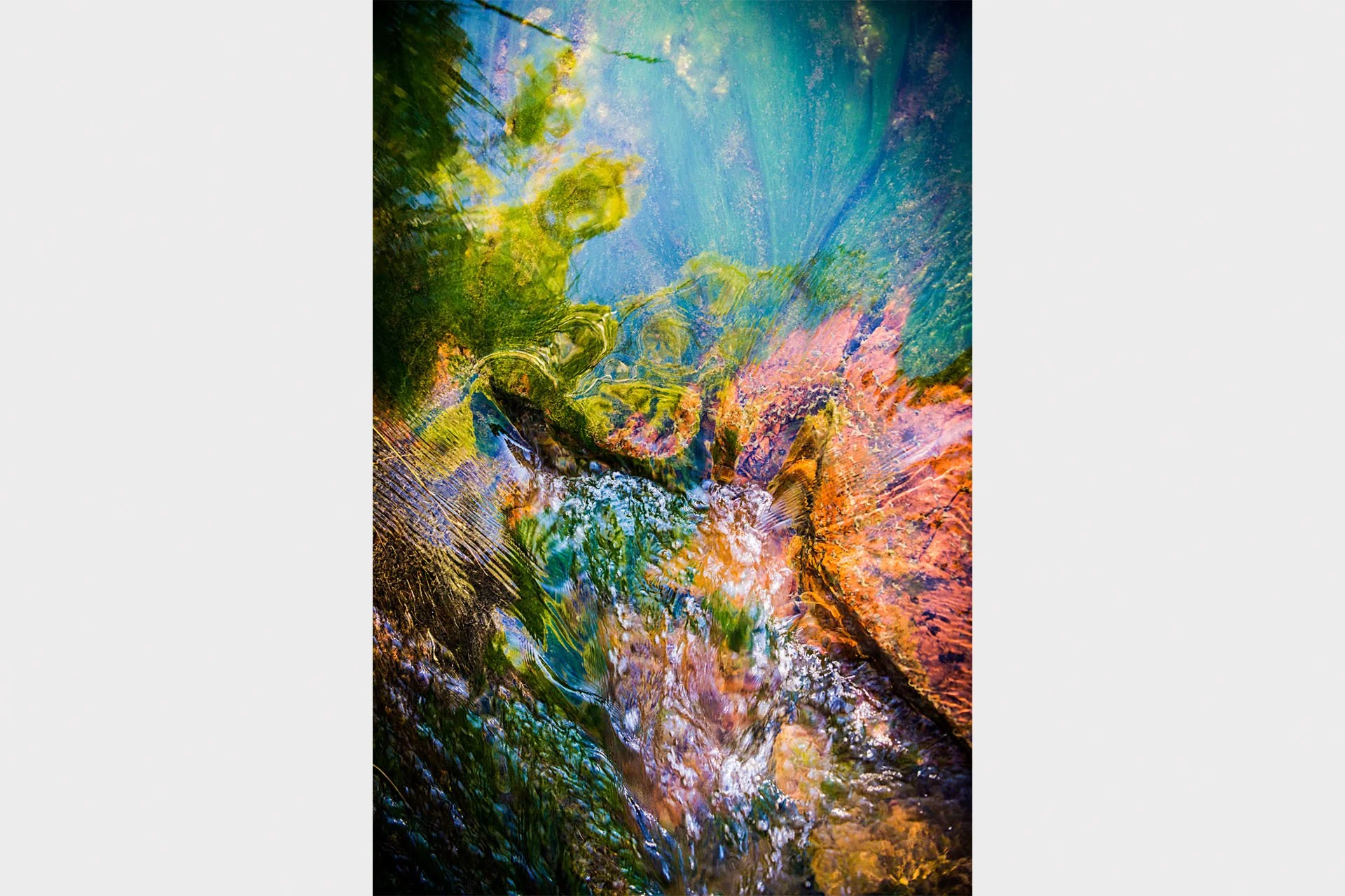Abstract close-up of clear water rippling over red rocks and green algae in a stream at Karijini National Park, Western Australia, reflecting blue sky in swirls of colour.