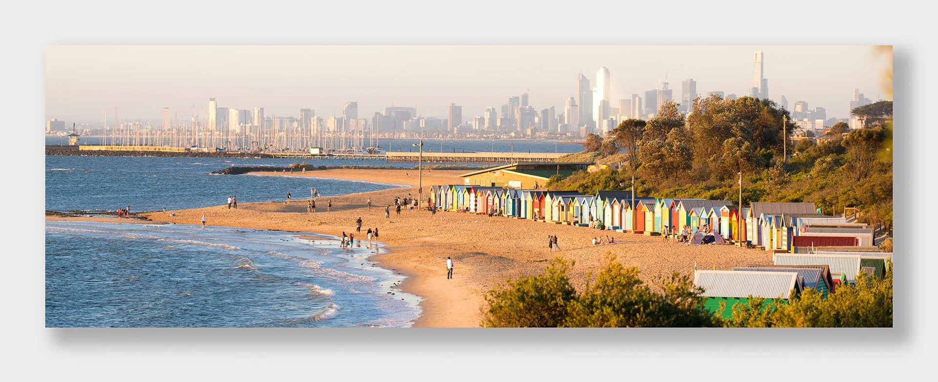 Panorama of Brighton Beach, Melbourne, Victoria, in warm late light, with colourful bathing boxes along the sand, people strolling the shore and the city skyline across the bay.