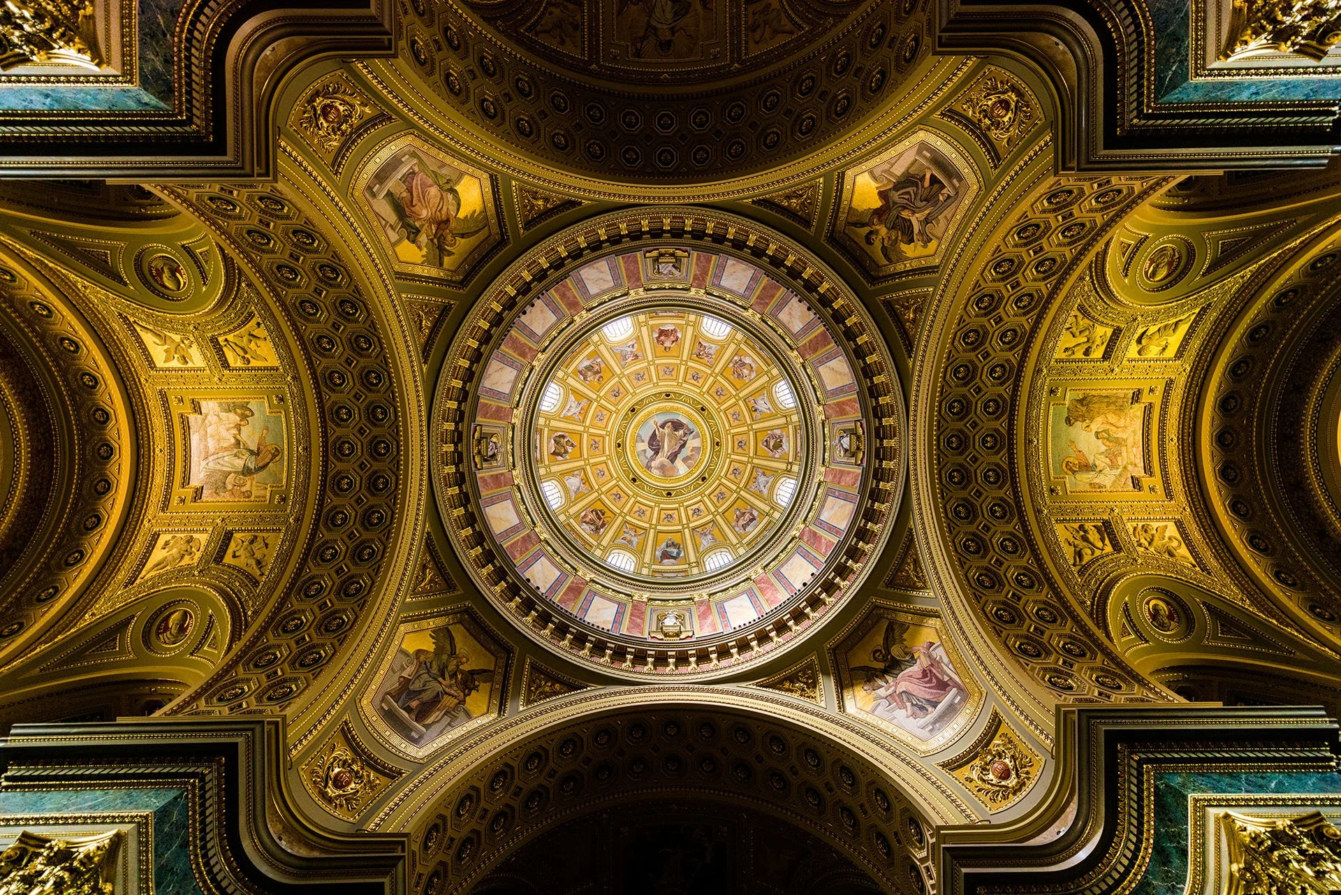 Looking straight up into the ornate gilded dome of St Stephen’s Basilica, Budapest, Hungary, with painted panels, coffered arches and a central mosaic ringed by windows.