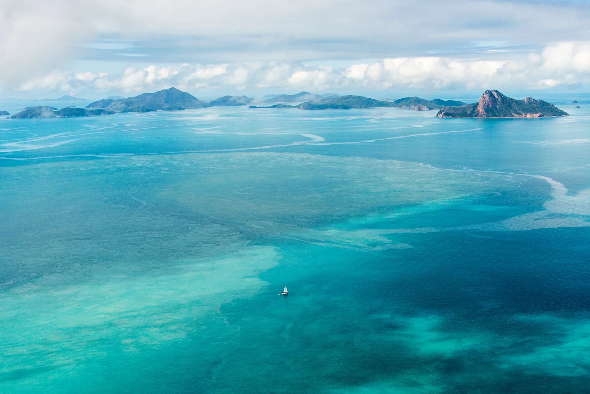 Aerial view of a lone sailing boat on turquoise water among the Whitsunday Islands, Queensland, with rugged green islands and clouds on the horizon.