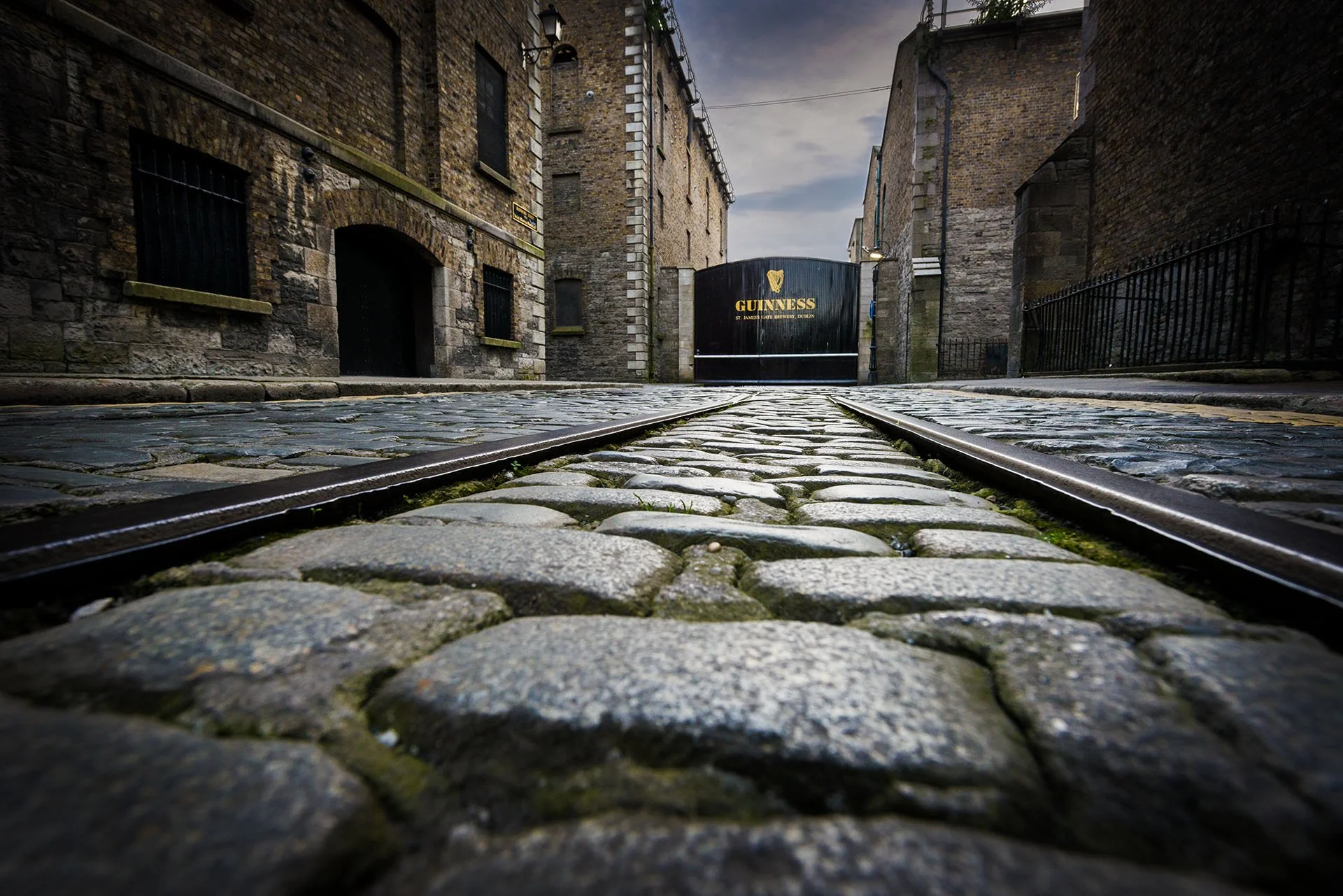 Low-angle view along old rail tracks set in cobblestones, leading between stone warehouses to the Guinness gate at St James’s Gate Brewery, Dublin, Ireland.