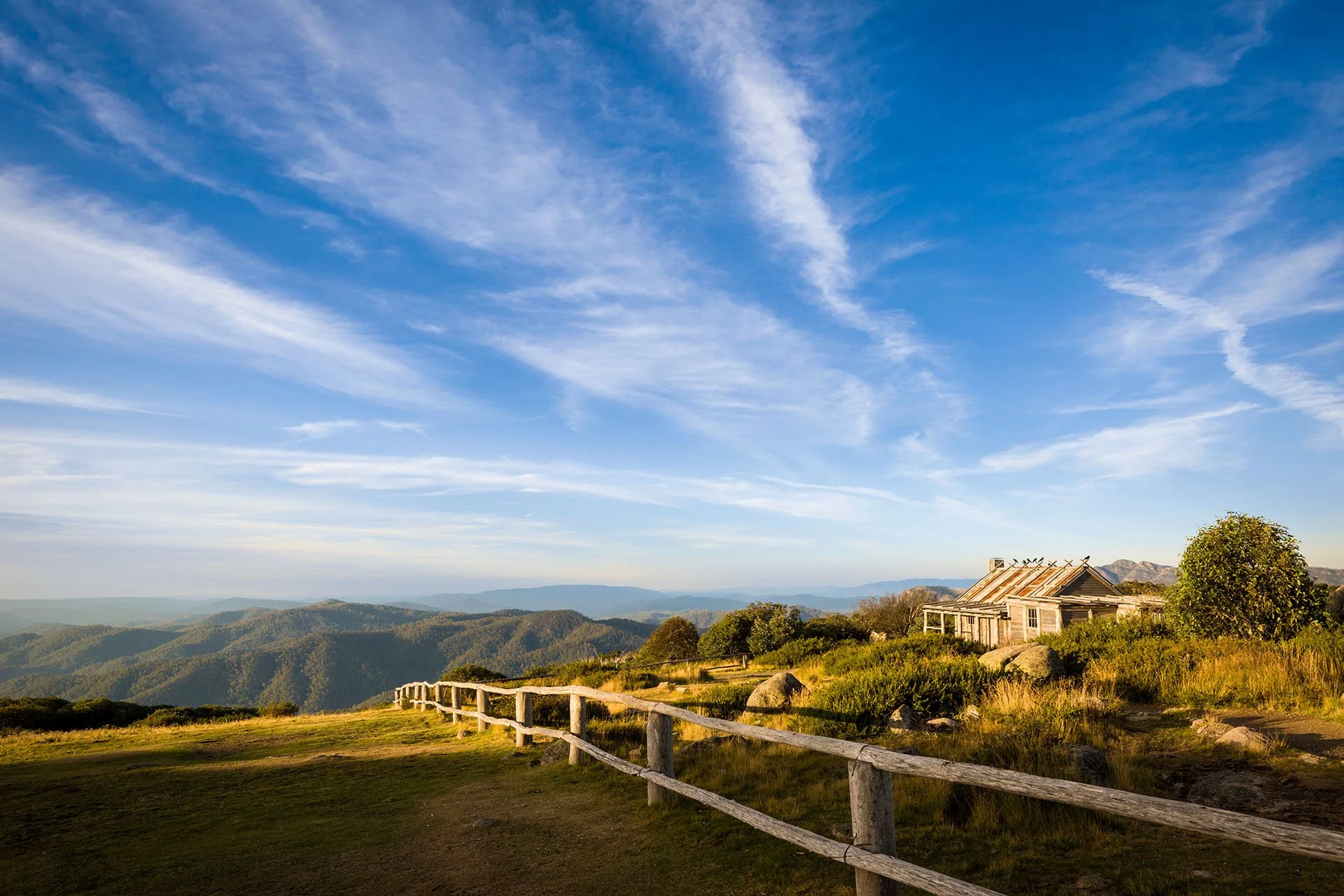 Craig’s Hut near Mount Stirling in the Victorian High Country, glowing in late afternoon light beside a rustic timber fence, with forested ranges and wispy clouds across a blue sky.