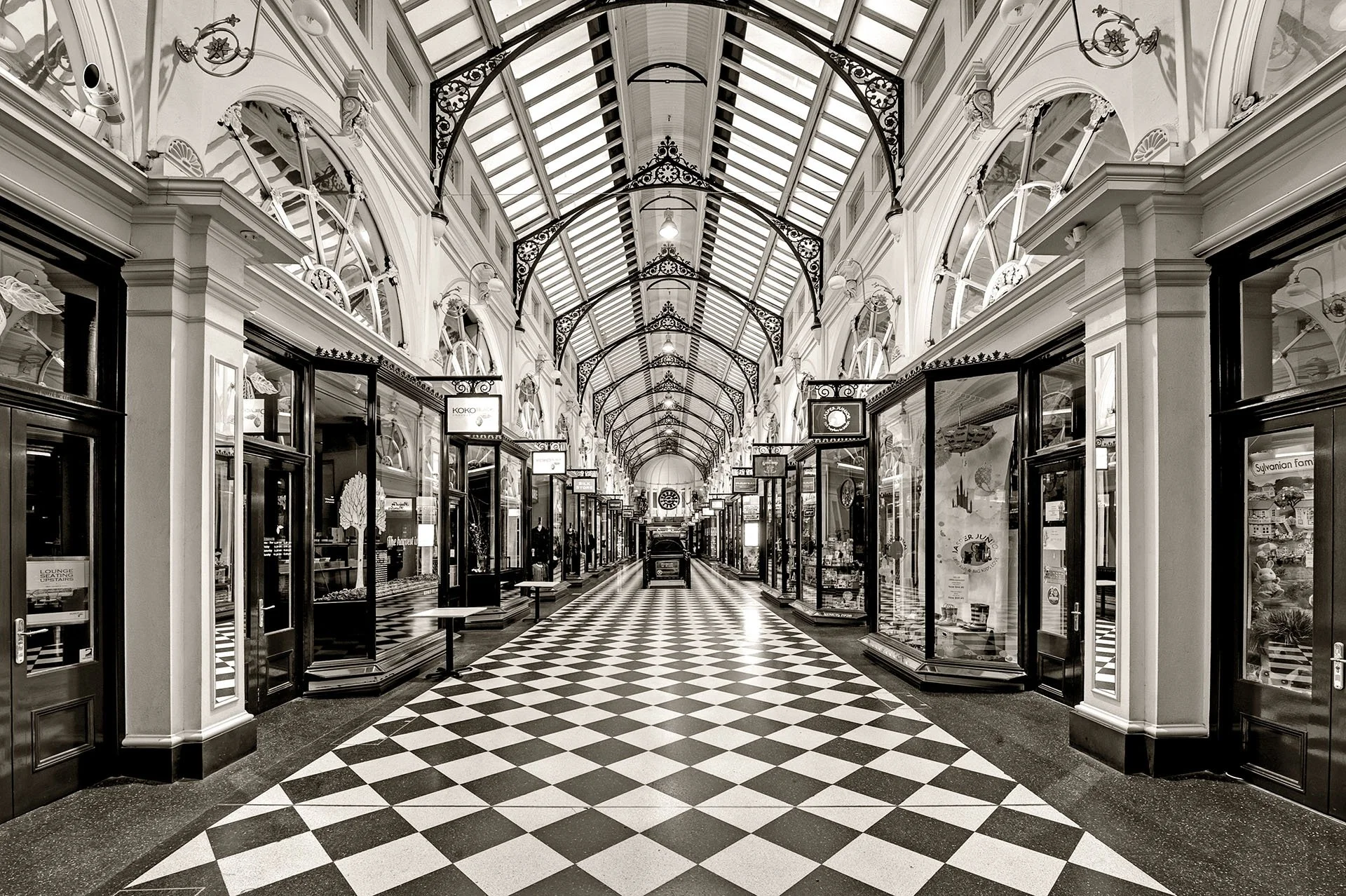 Black and white photo of the empty Royal Arcade in Melbourne, Victoria, with a chequered tile floor leading between shopfronts under an arched glass roof with ornate iron trusses.