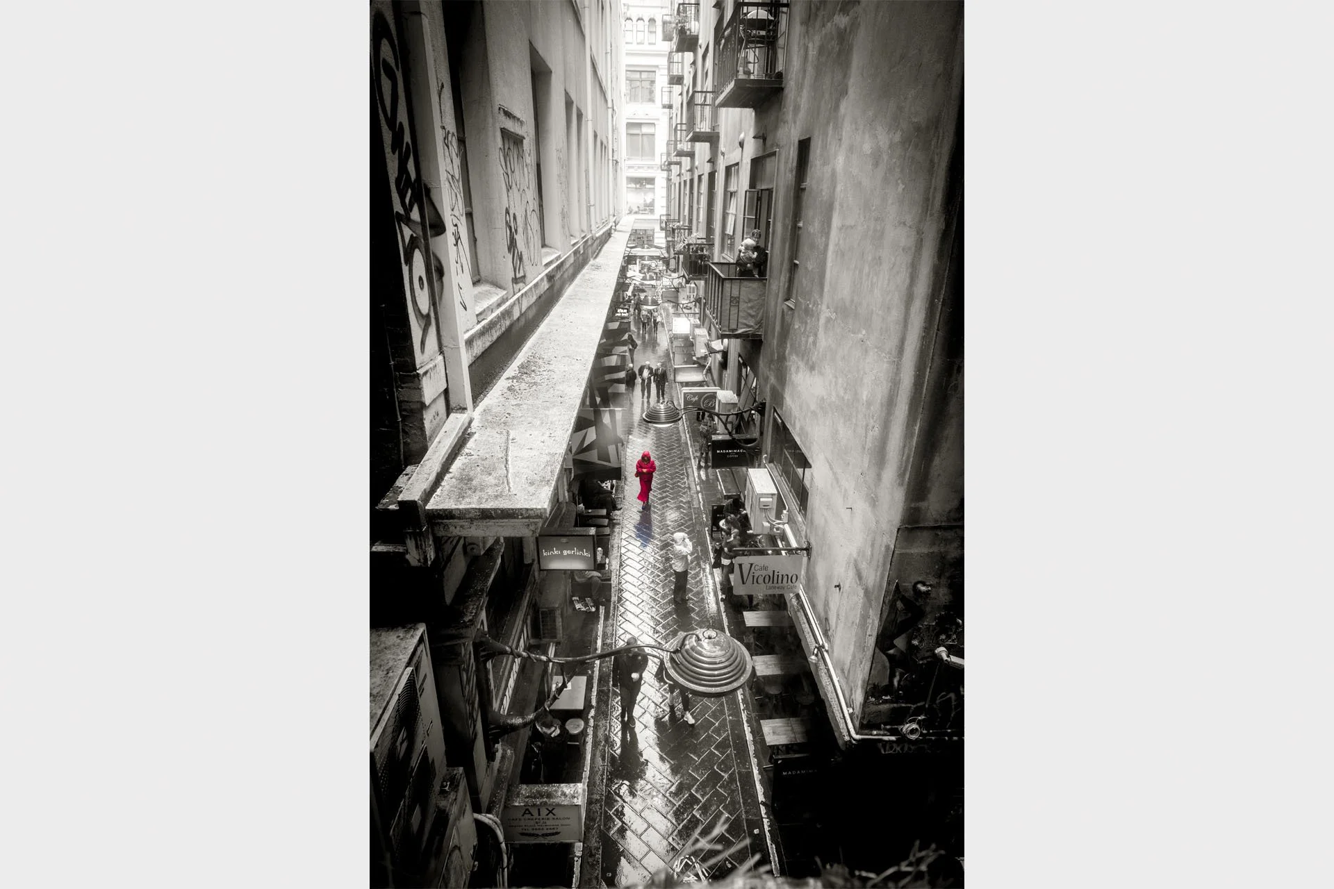 Black and white view looking down into Centre Place laneway in Melbourne on a wet day, with a figure in a red coat walking along the cobblestones between cafes and graffiti-covered walls.